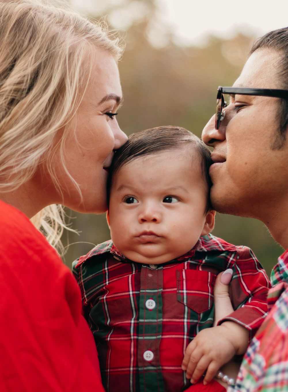 Sweet family portrait showing parents kissing baby in plaid outfit outdoors.