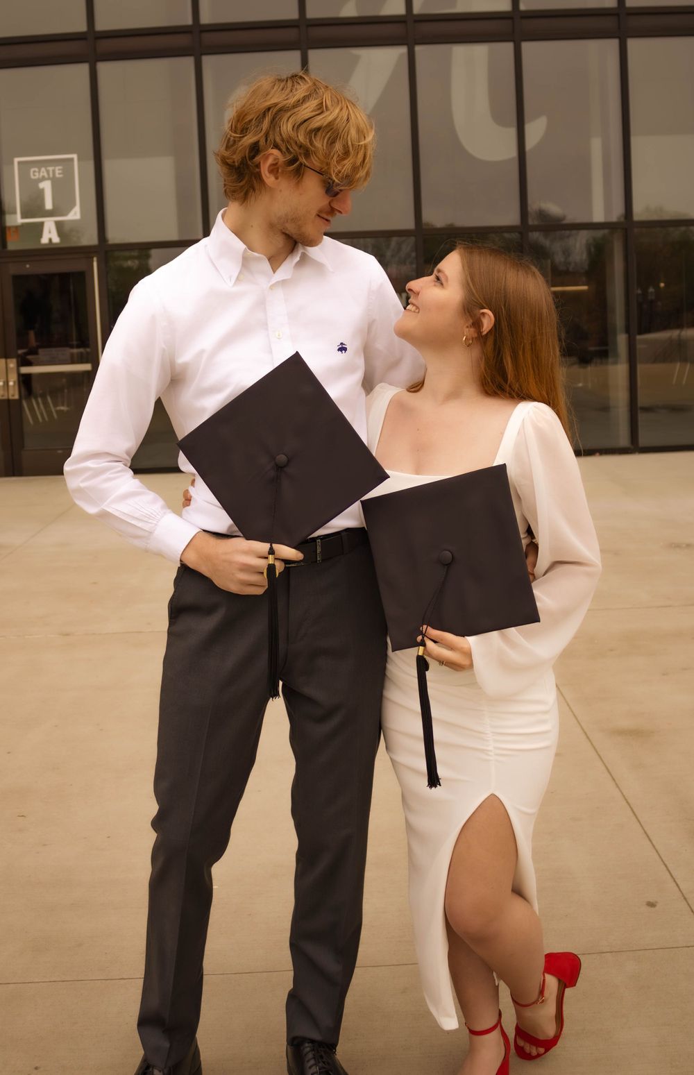 Graduation couple in formal attire holding diplomas outside modern building.