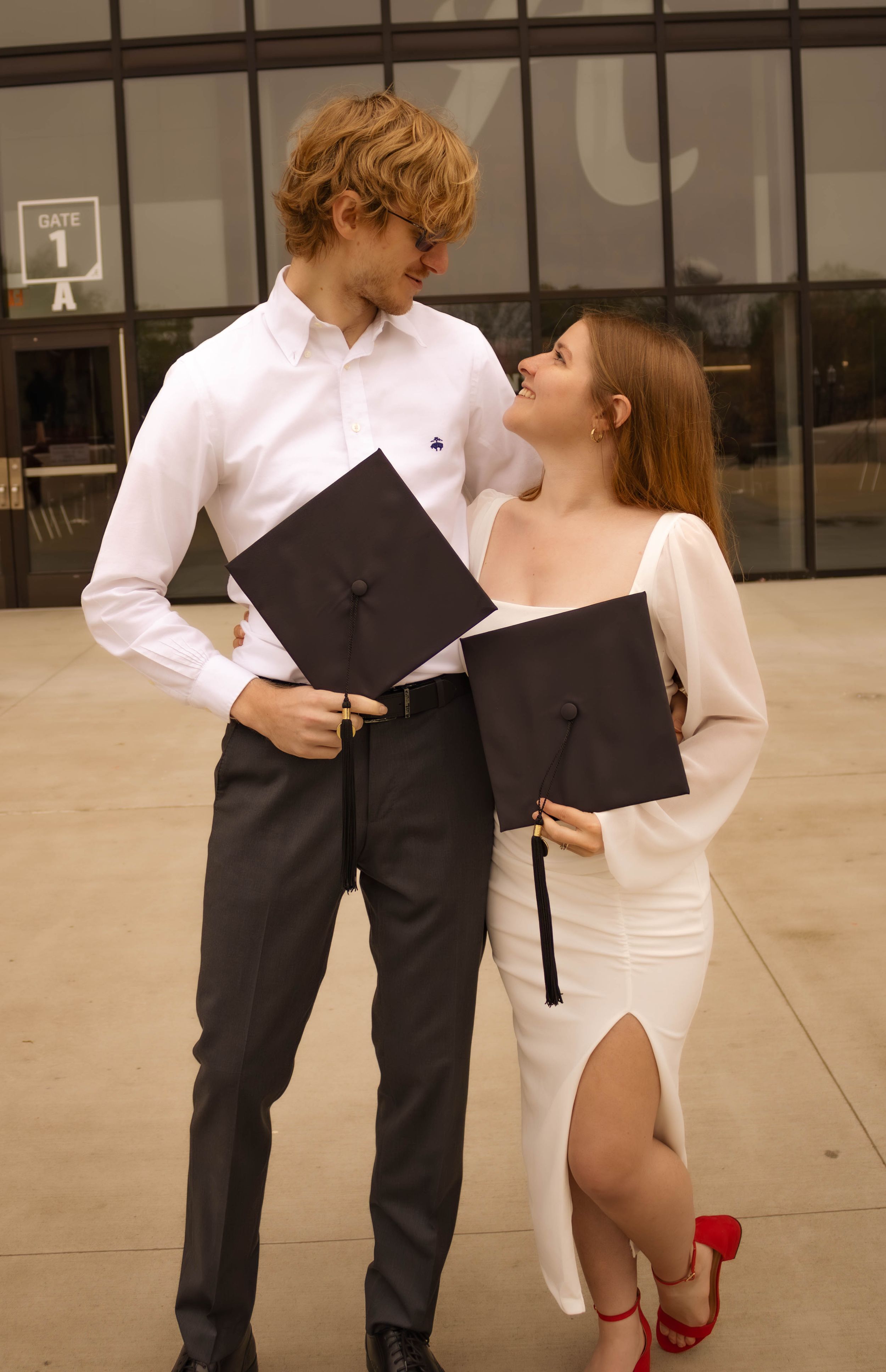 Graduation couple in formal attire holding diplomas outside modern building.
