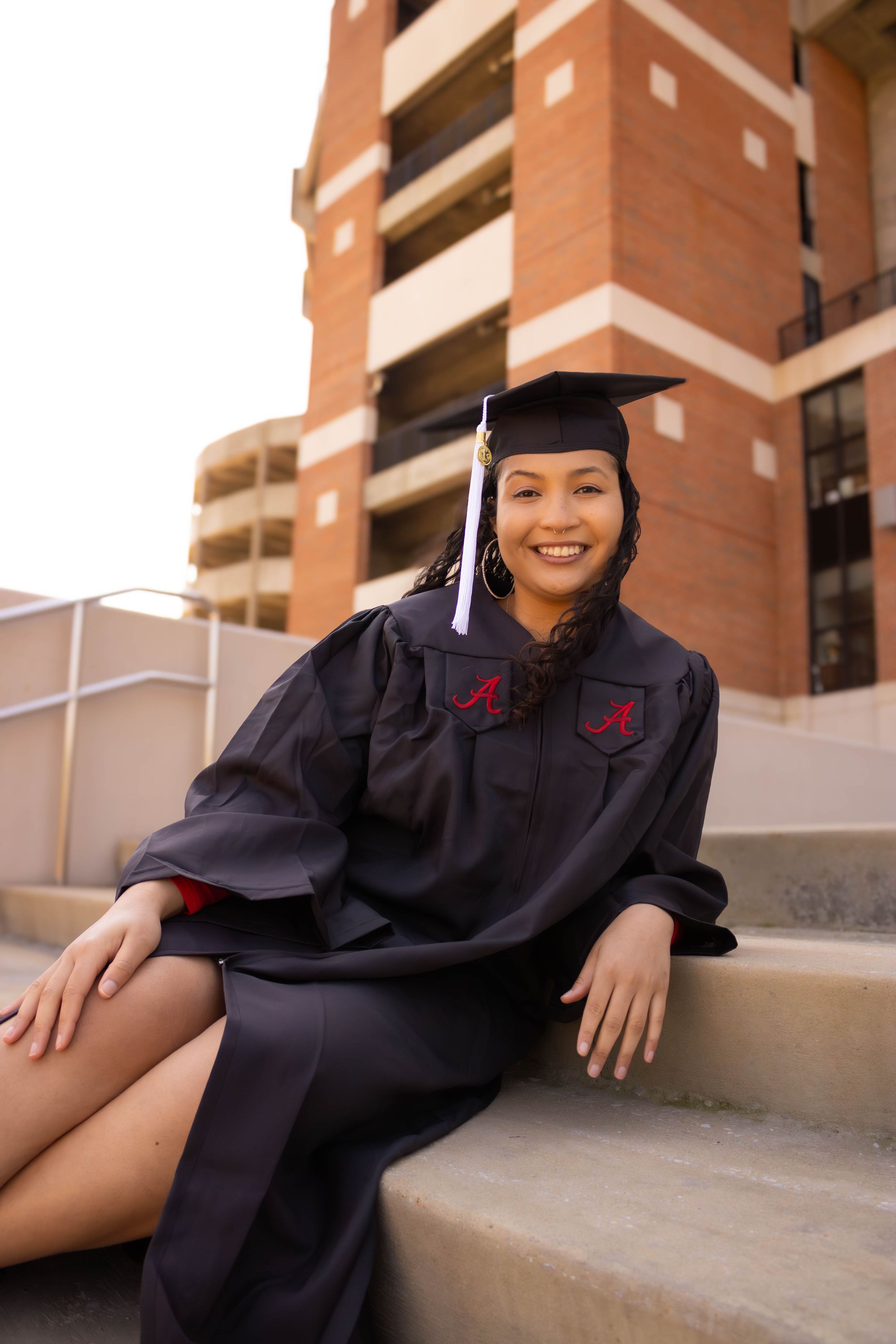 Graduate in black cap and gown sitting on concrete steps near brick building.