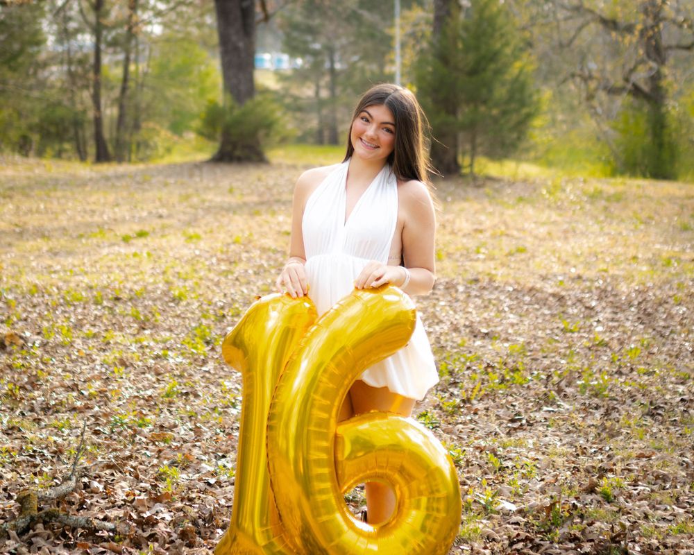 Person in white dress poses with large gold number 6 balloon in autumn setting.