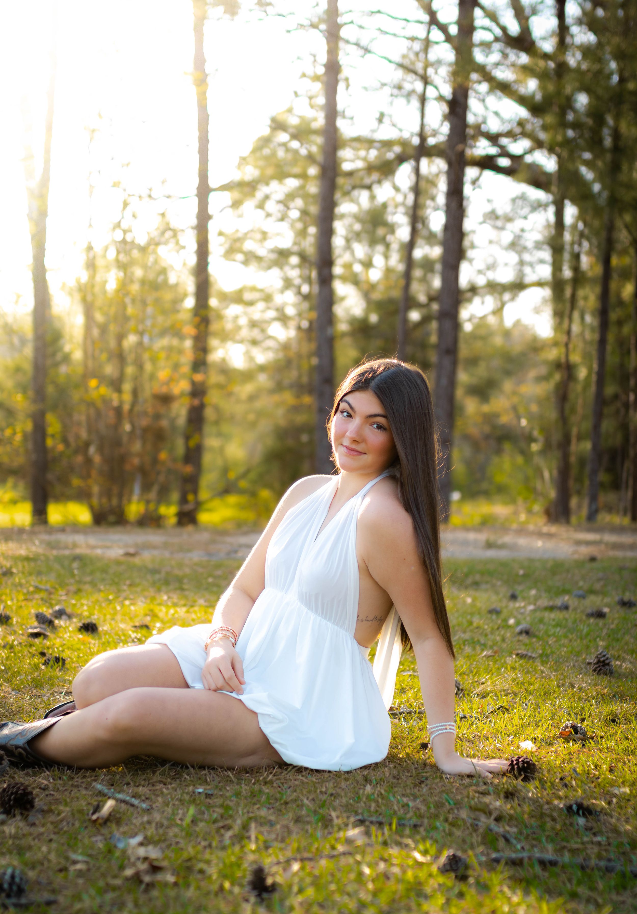 Person in white sundress sitting on grass in sunlit forest setting.