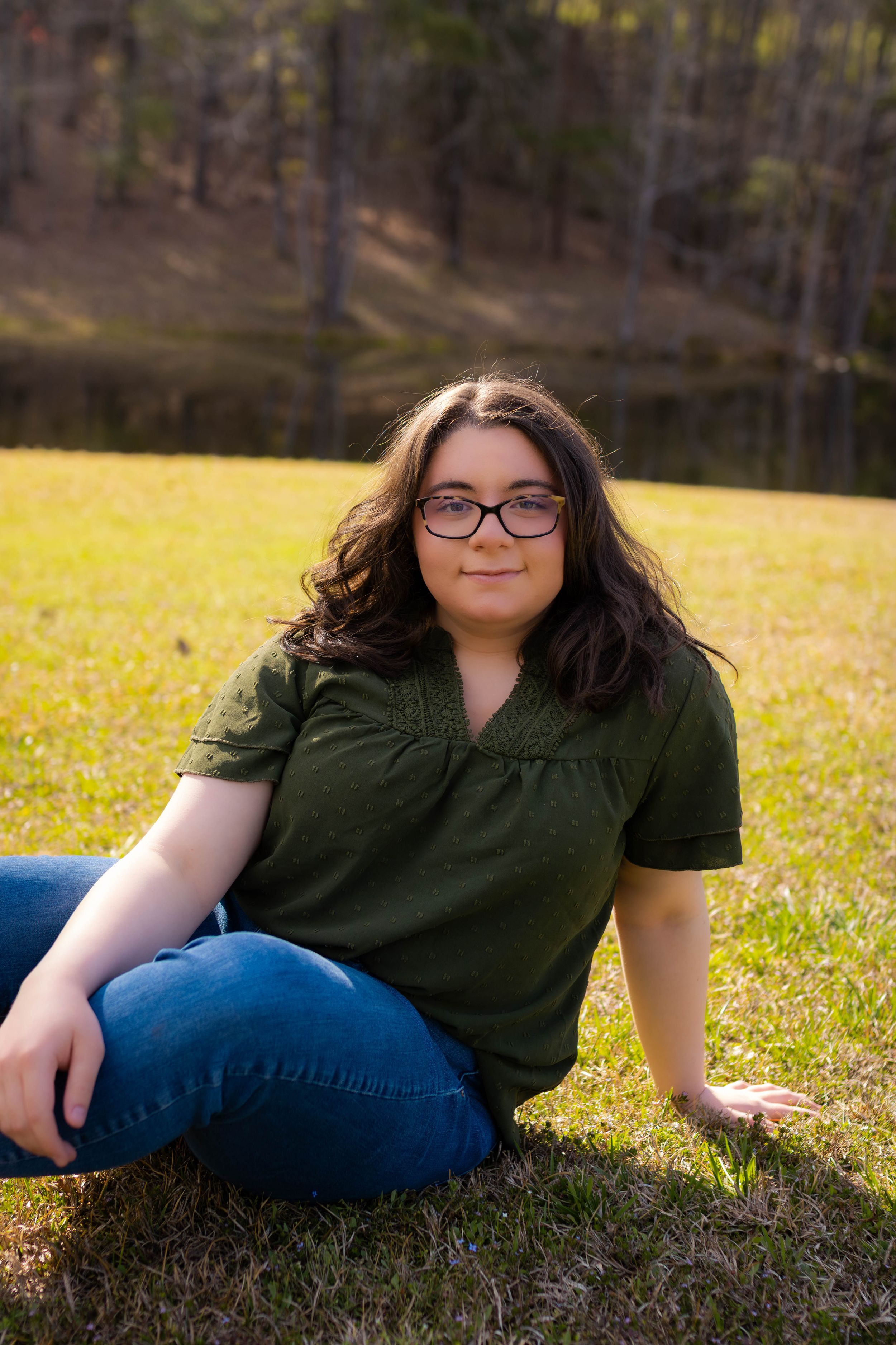 Outdoor portrait of person in green top and glasses sitting on grass near lake.