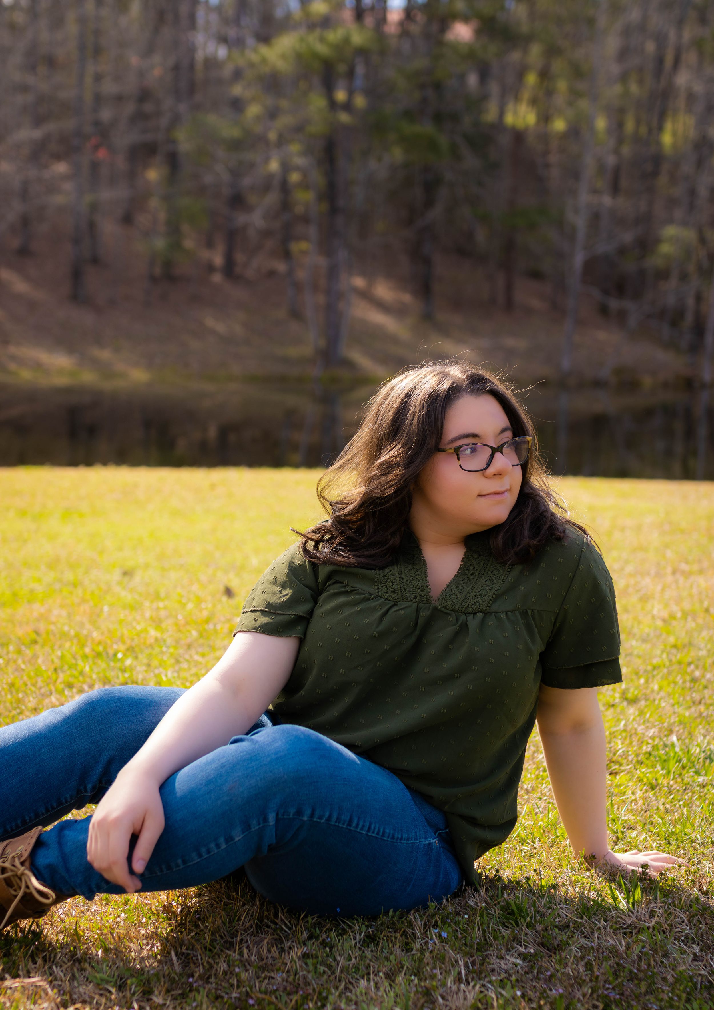 Casual portrait of someone in green shirt and jeans sitting on grassy lawn by lake.