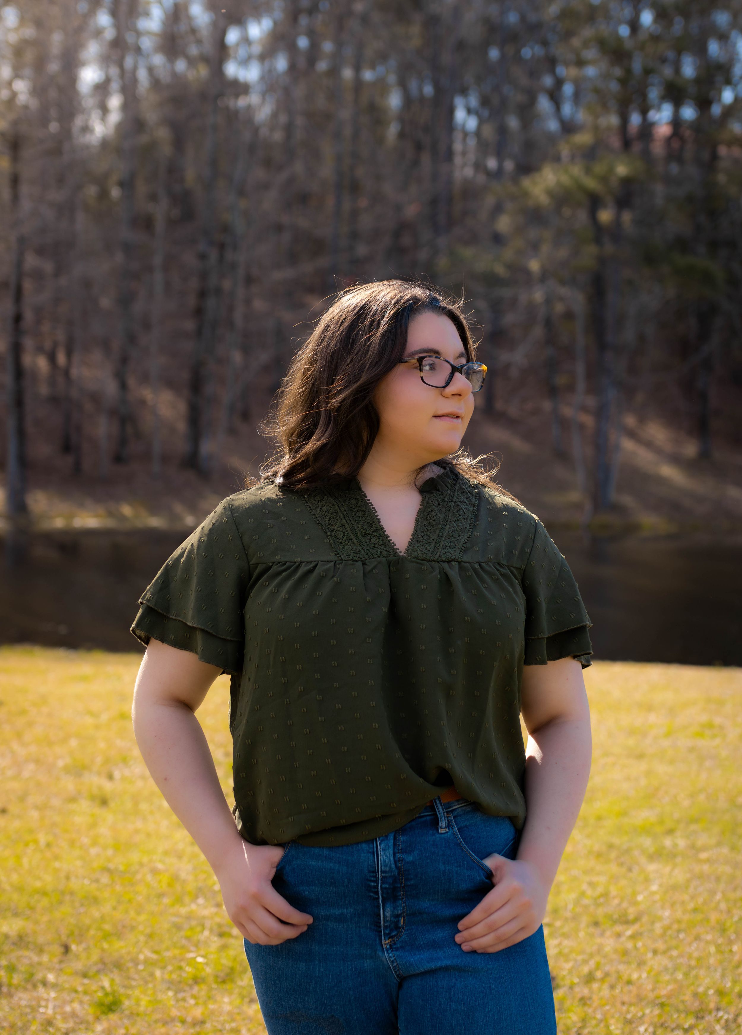 Casual outdoor photo of someone in olive green top and jeans standing by lake.