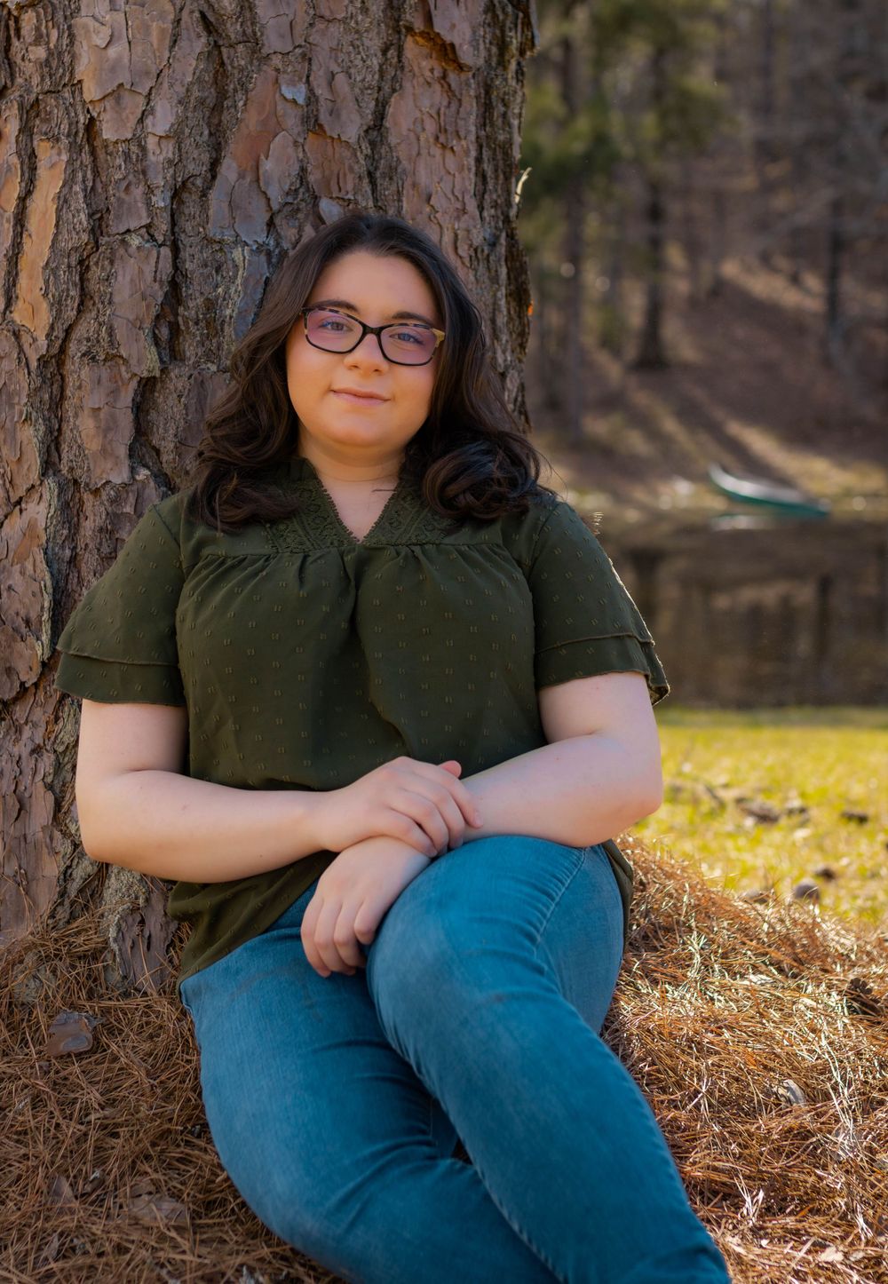Portrait of person in dark green top leaning against pine tree trunk by lake.