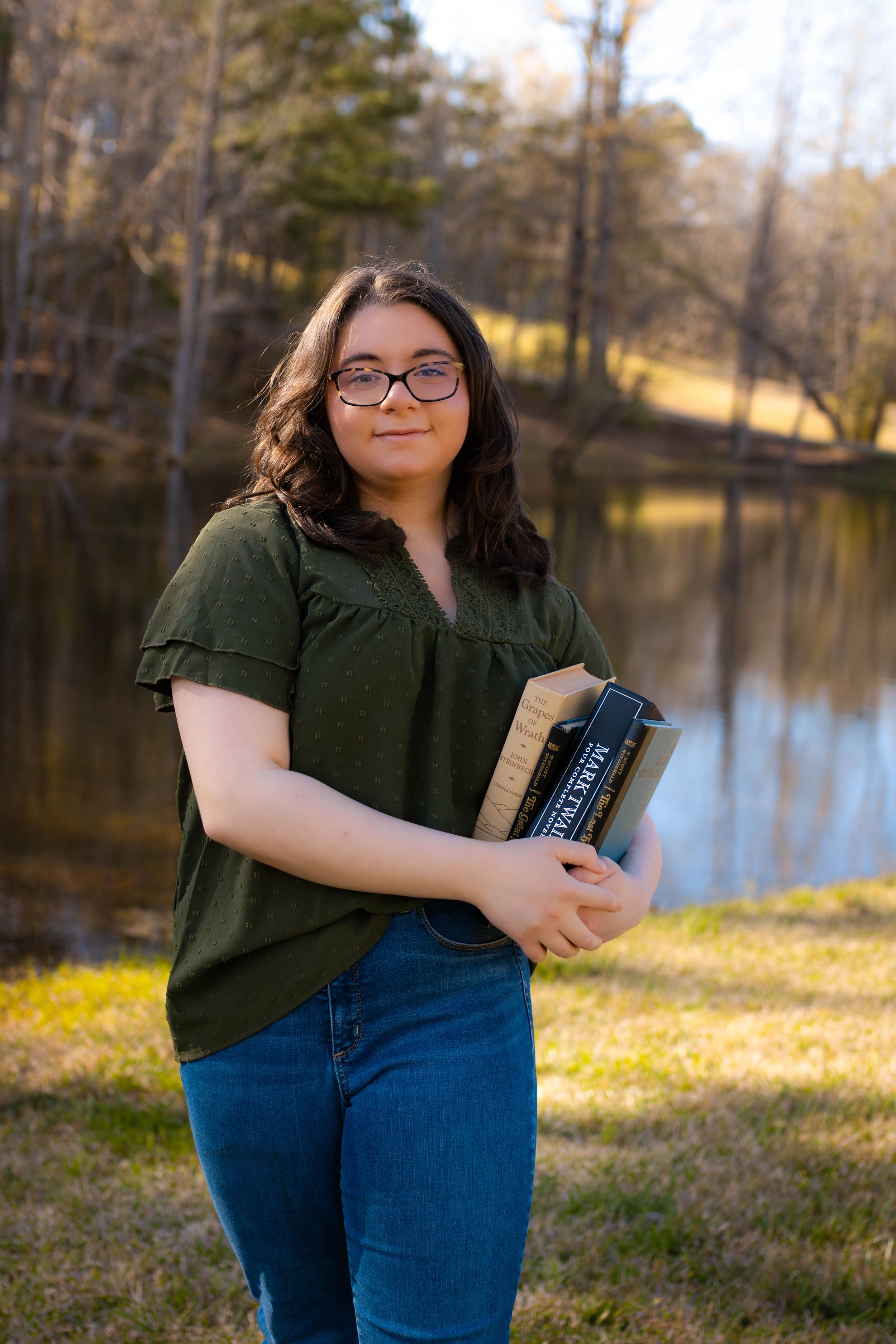 Student in olive green top holds book while standing near lakeside in wooded setting.