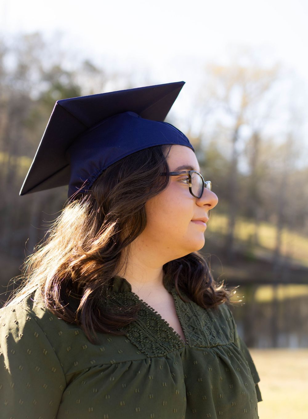 Graduate in a dark green dress and blue cap stands outdoors on a sunny day.