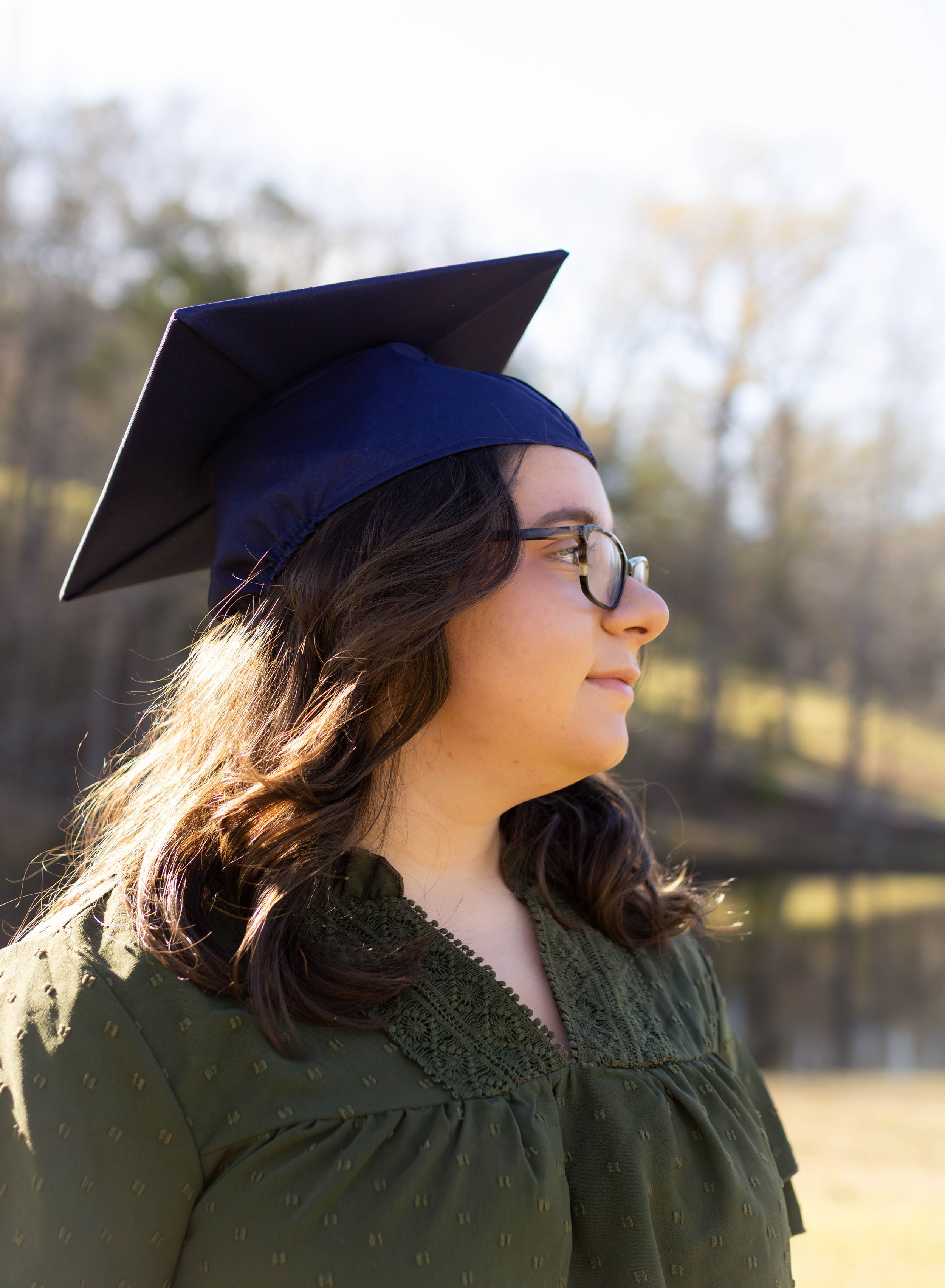 Graduate in a dark green dress and blue cap stands outdoors on a sunny day.