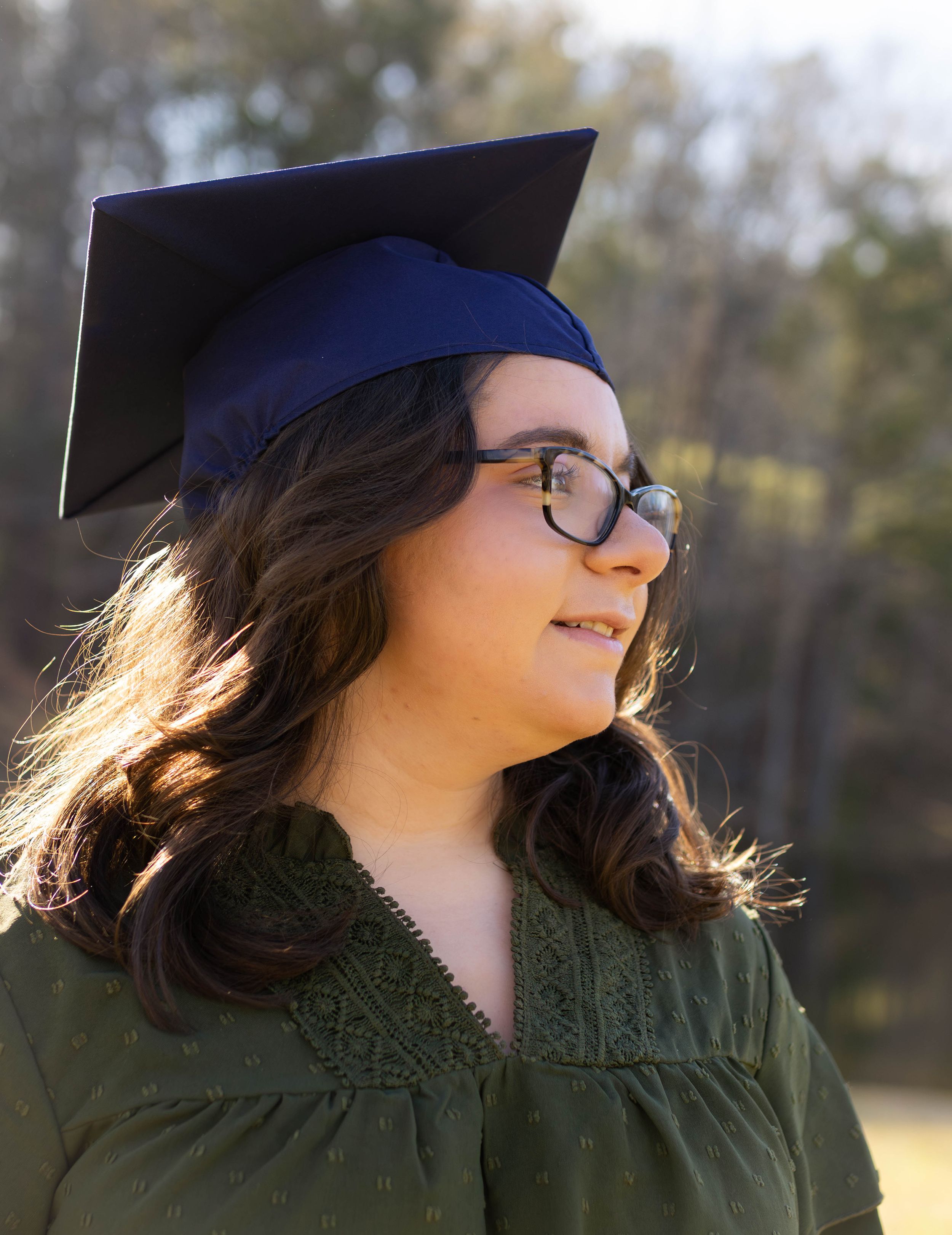 Graduate in dark green dress and cap poses outdoors with natural lighting.