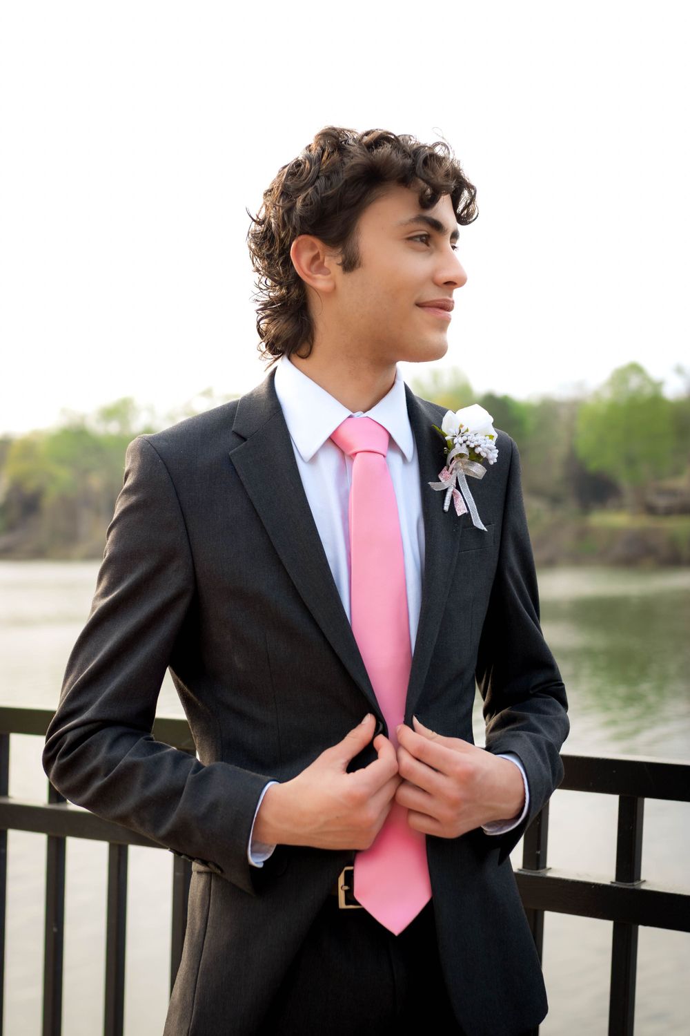 Young man in black suit with pink tie and boutonniere poses by waterfront railing.