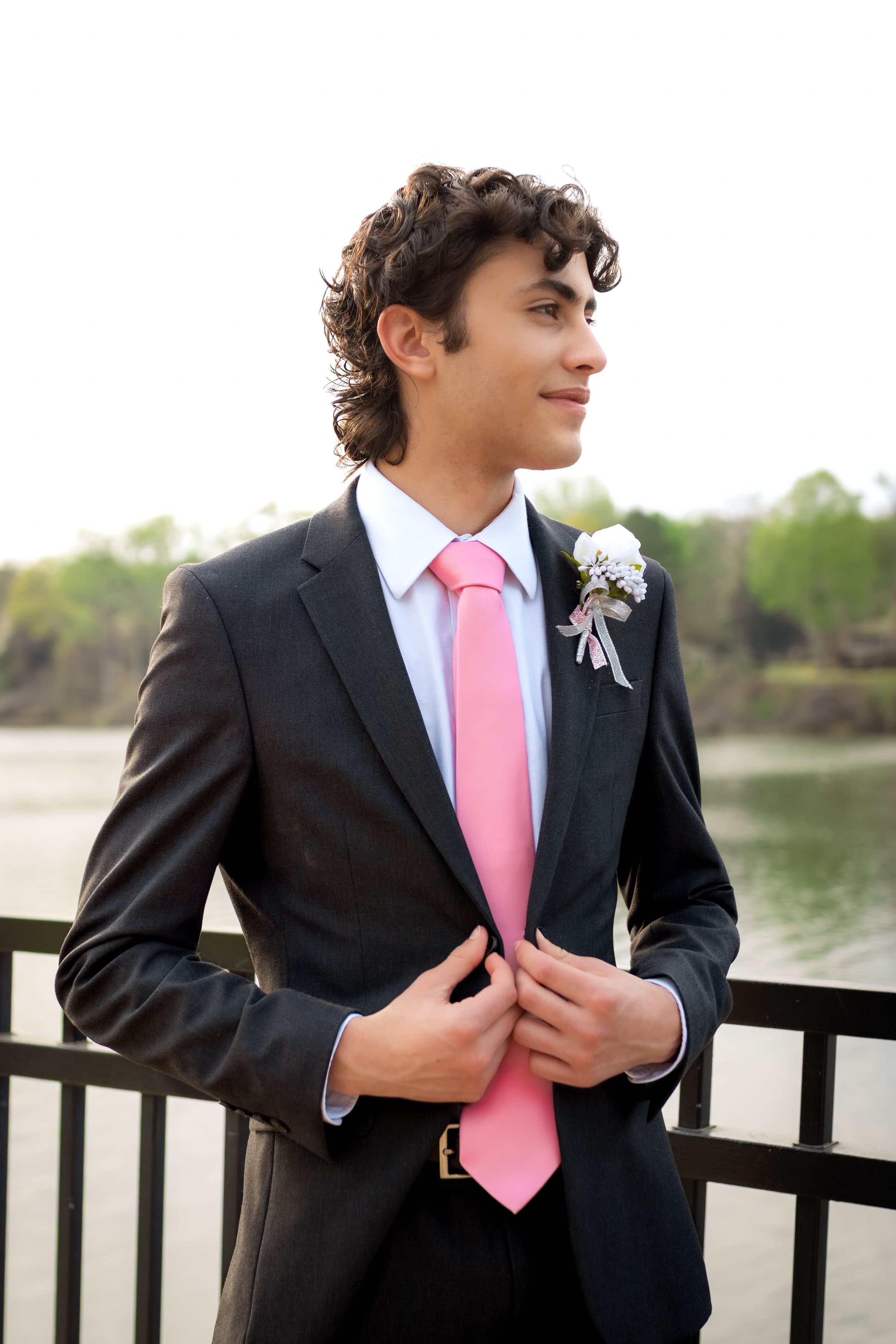 Young man in black suit with pink tie and boutonniere poses by waterfront railing.