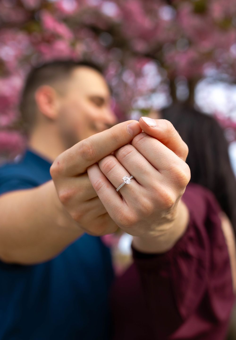 Close-up of hand showing engagement ring with cherry blossoms in background.