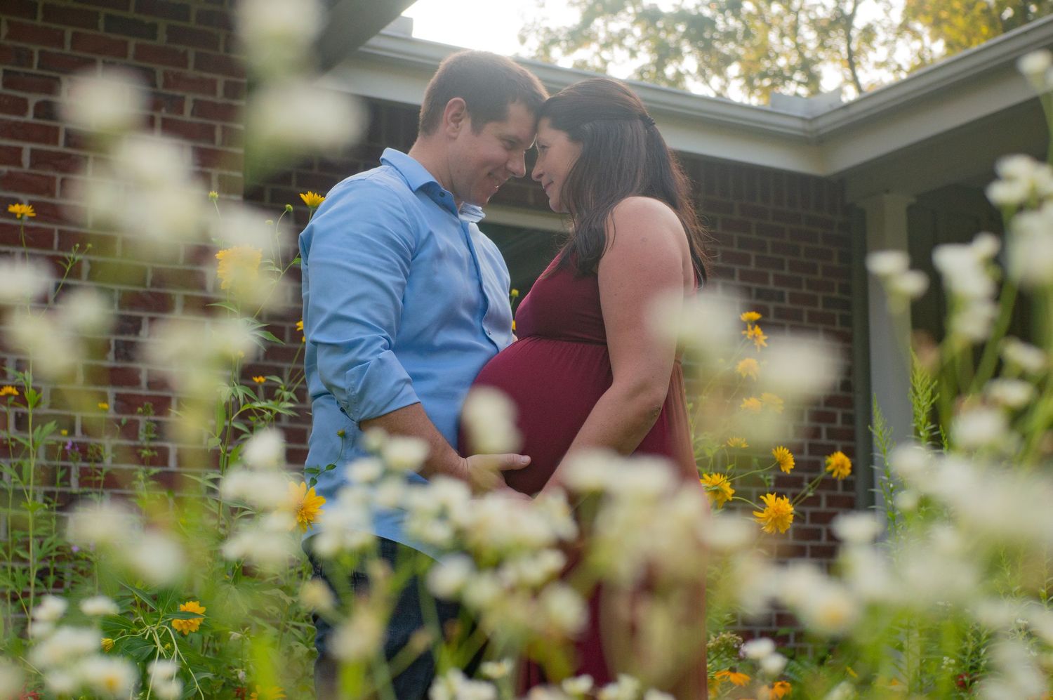 A romantic moment between soon-to-be parents captured through blooming white wildflowers in a garden setting outside their brick house.