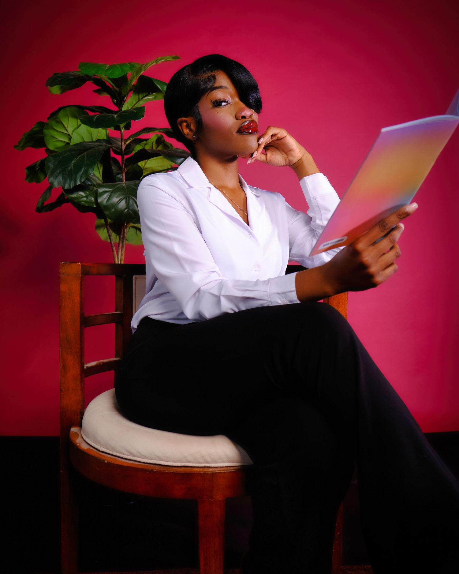 Professional in white blazer reading documents while seated near plant against pink backdrop.