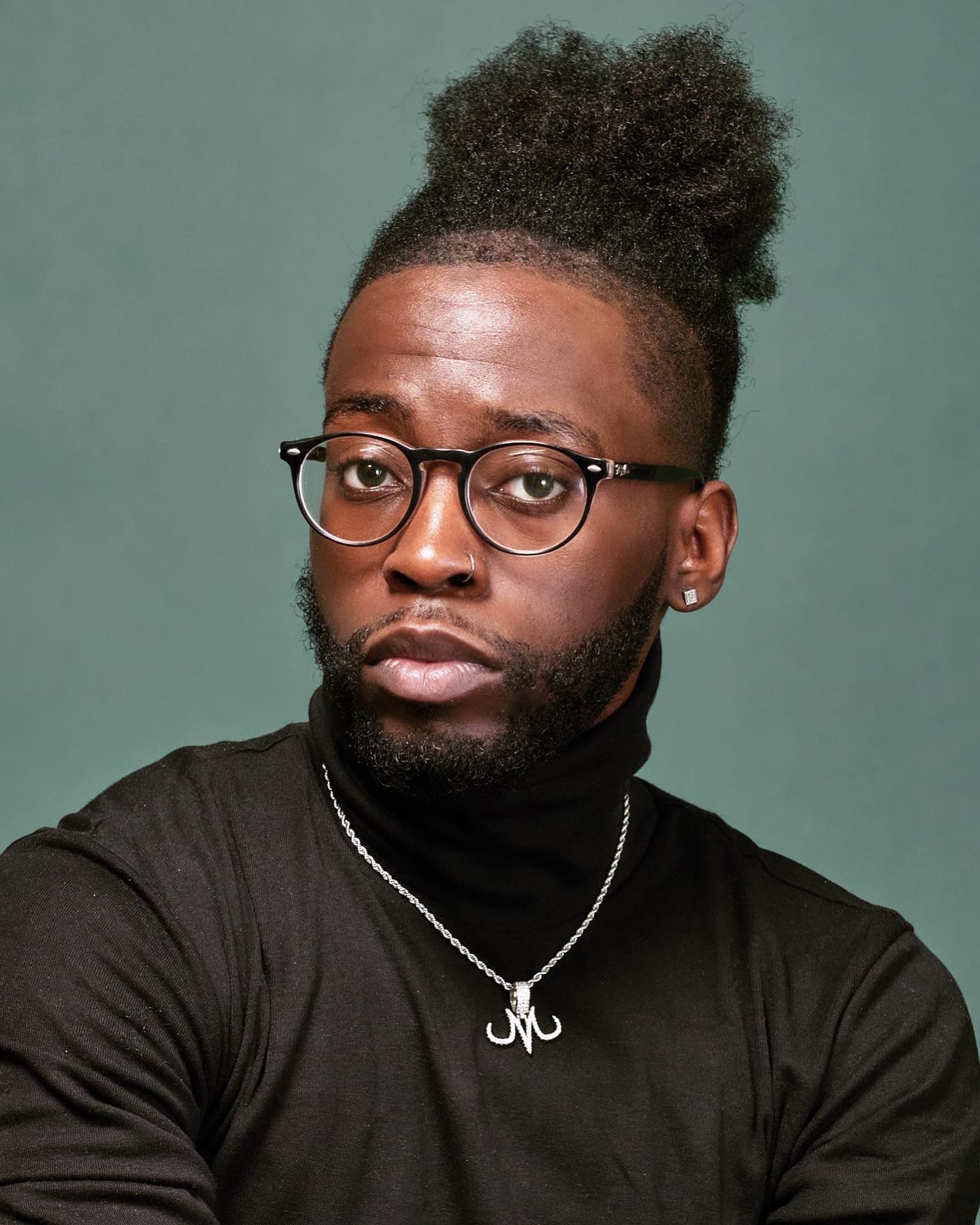 Studio portrait featuring glasses, top knot hairstyle, and black turtleneck against green background.