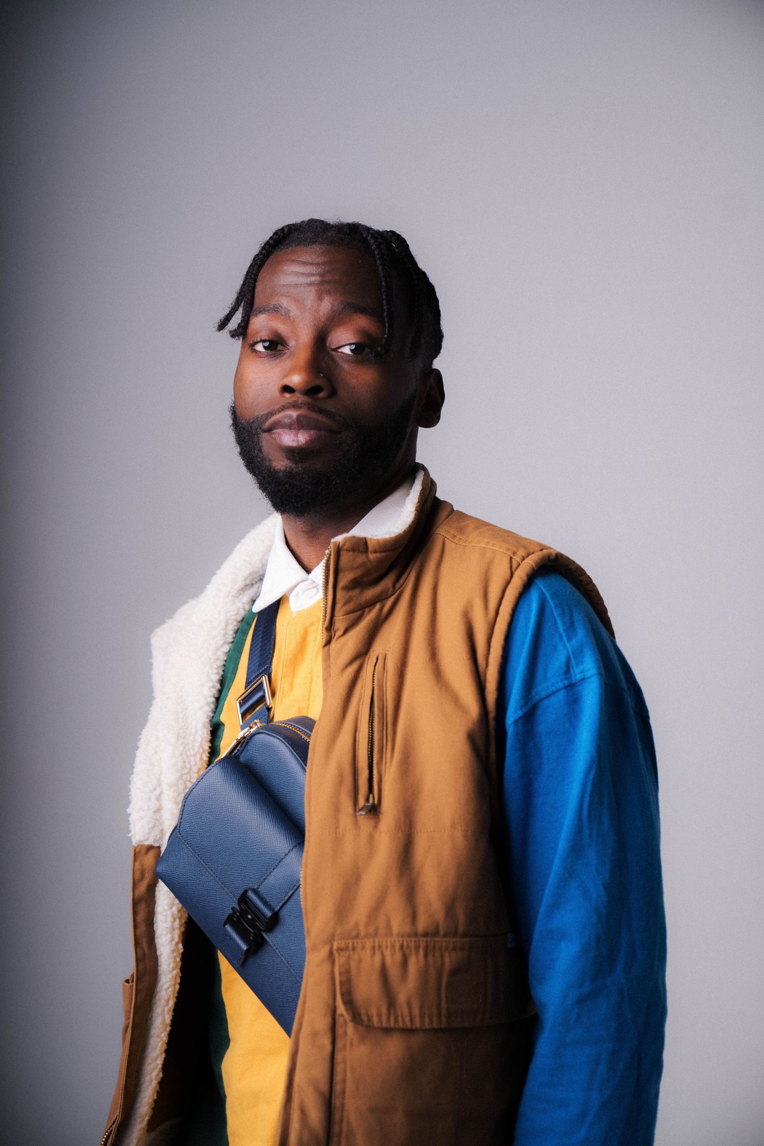 Portrait of person in color-block vest and blue sleeves holding navy bag against grey background.