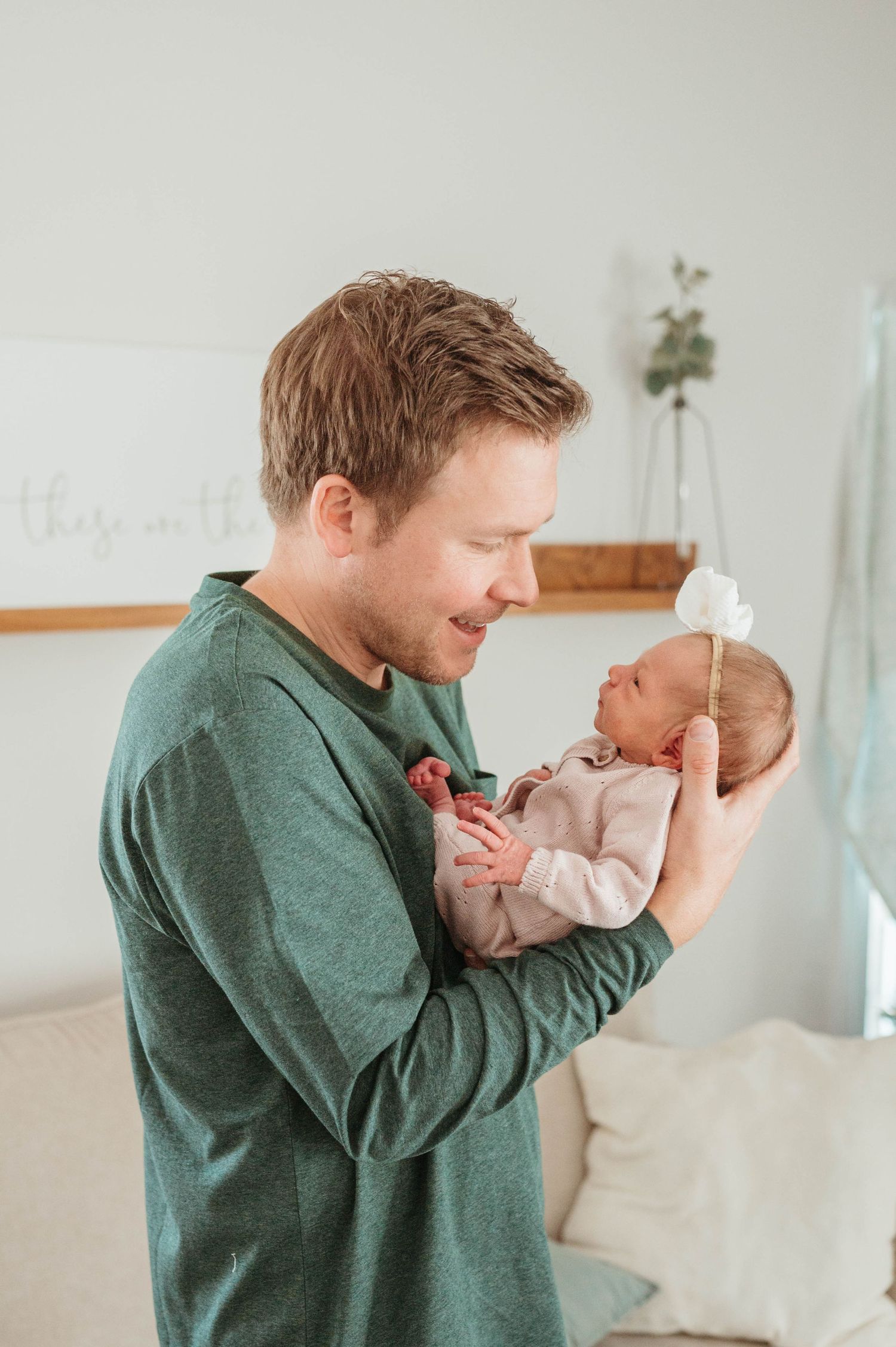 Loving father in green shirt gazes at his newborn baby girl wearing a delicate white bow headband.
