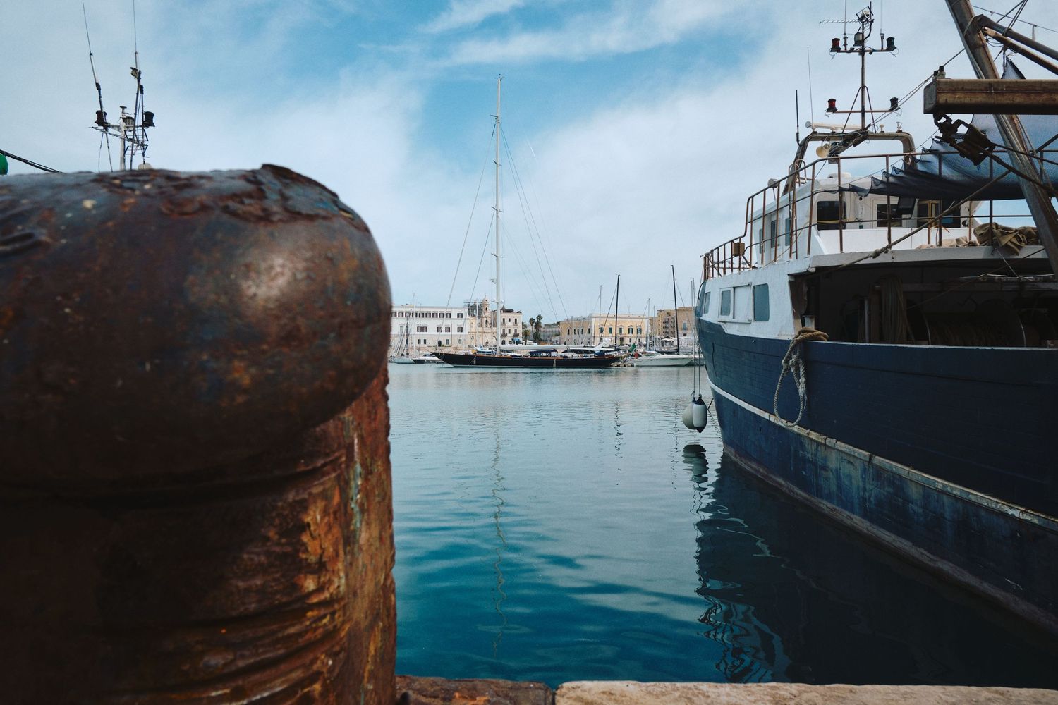 Rusty mooring bollard and blue fishing boat in a peaceful harbor with calm waters.