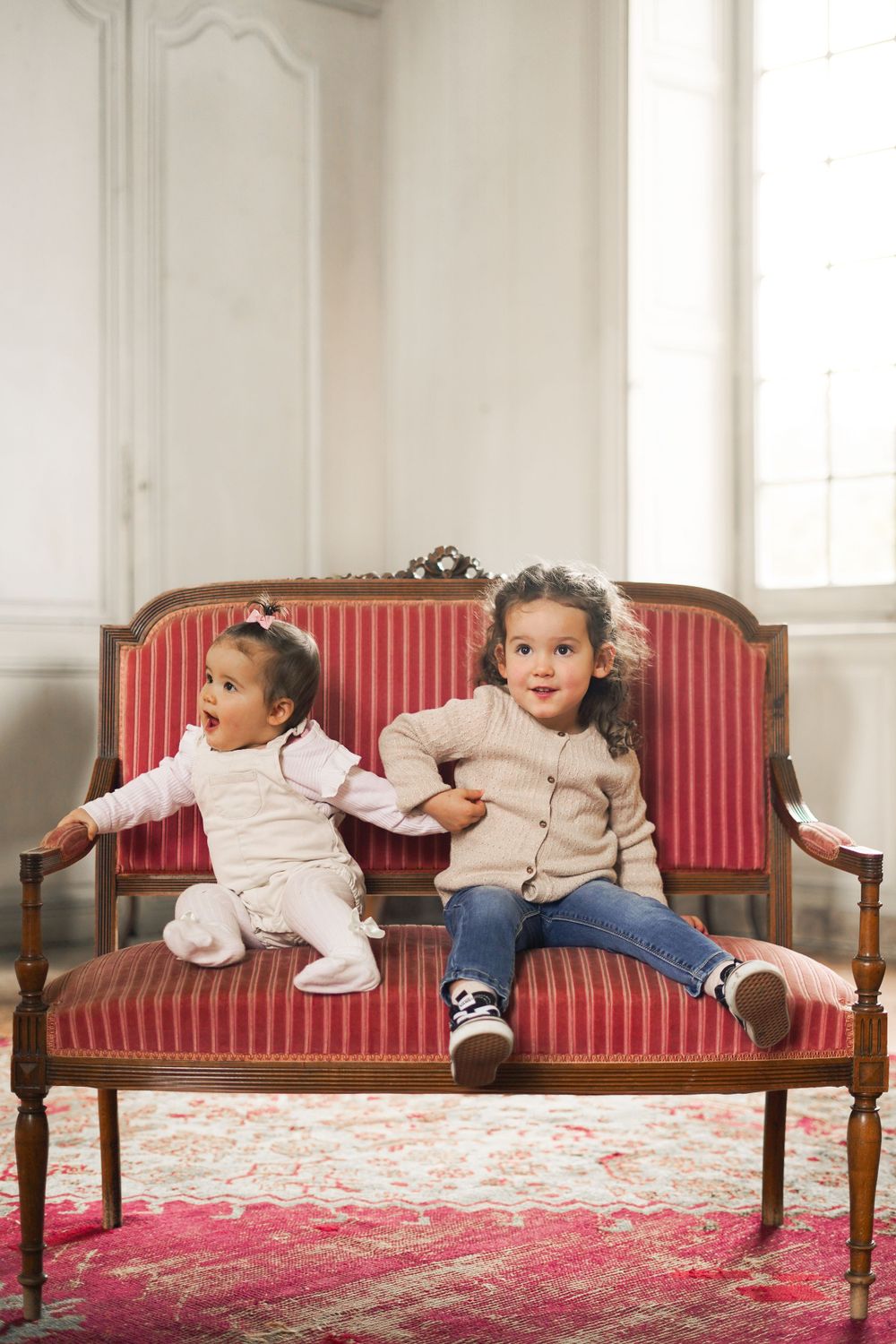 Two children sitting on vintage red striped settee in bright white room.