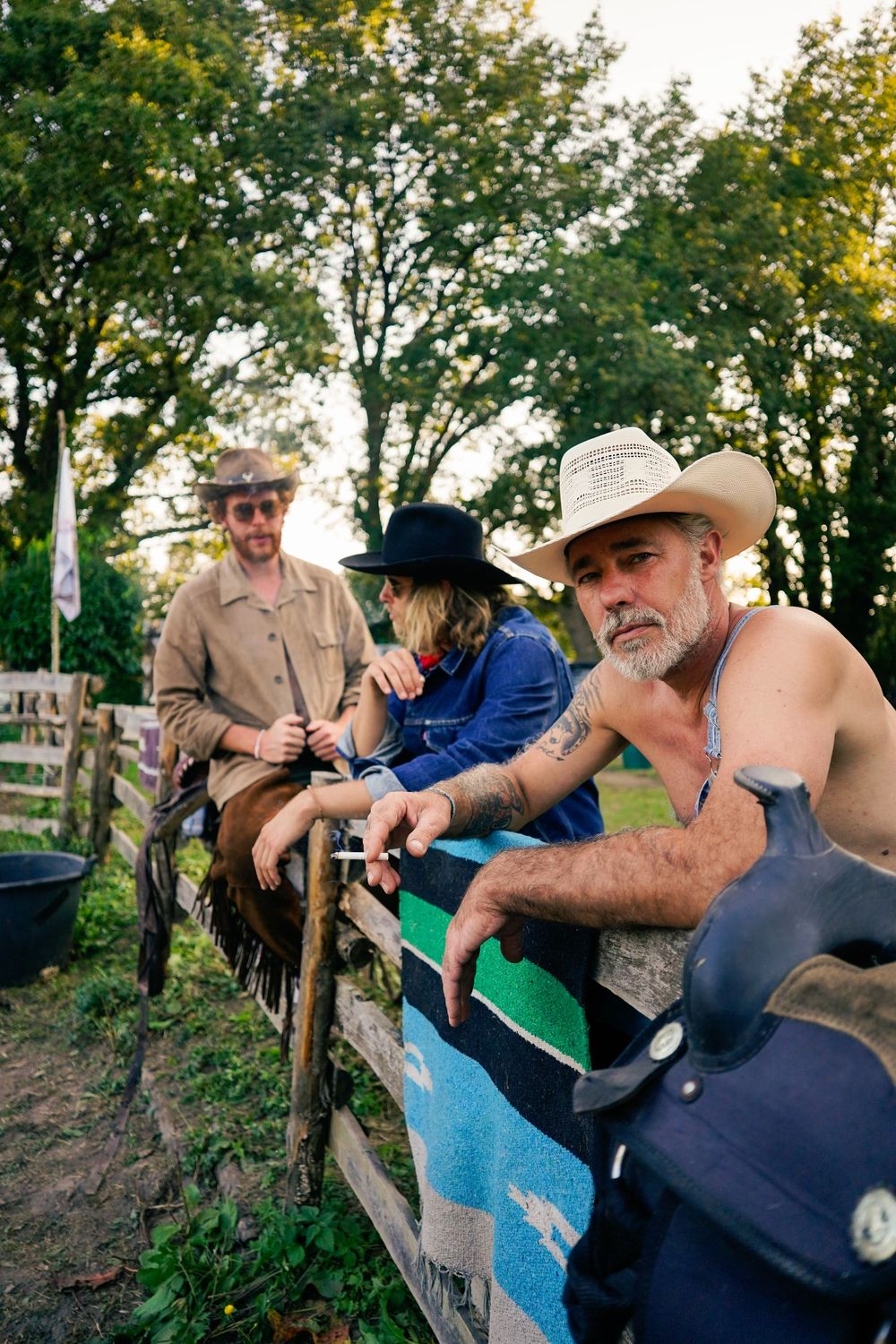 Three individuals in cowboy hats lean against a rustic wooden fence at a rural ranch setting.