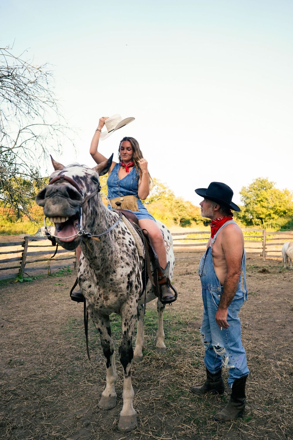 A rider atop a spotted horse waves their hat while someone in denim overalls stands nearby in a paddock.
