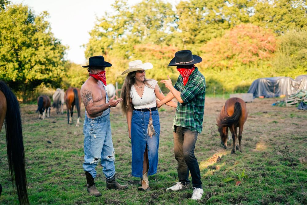 Three people dancing together in casual clothes in a field with horses in the background.