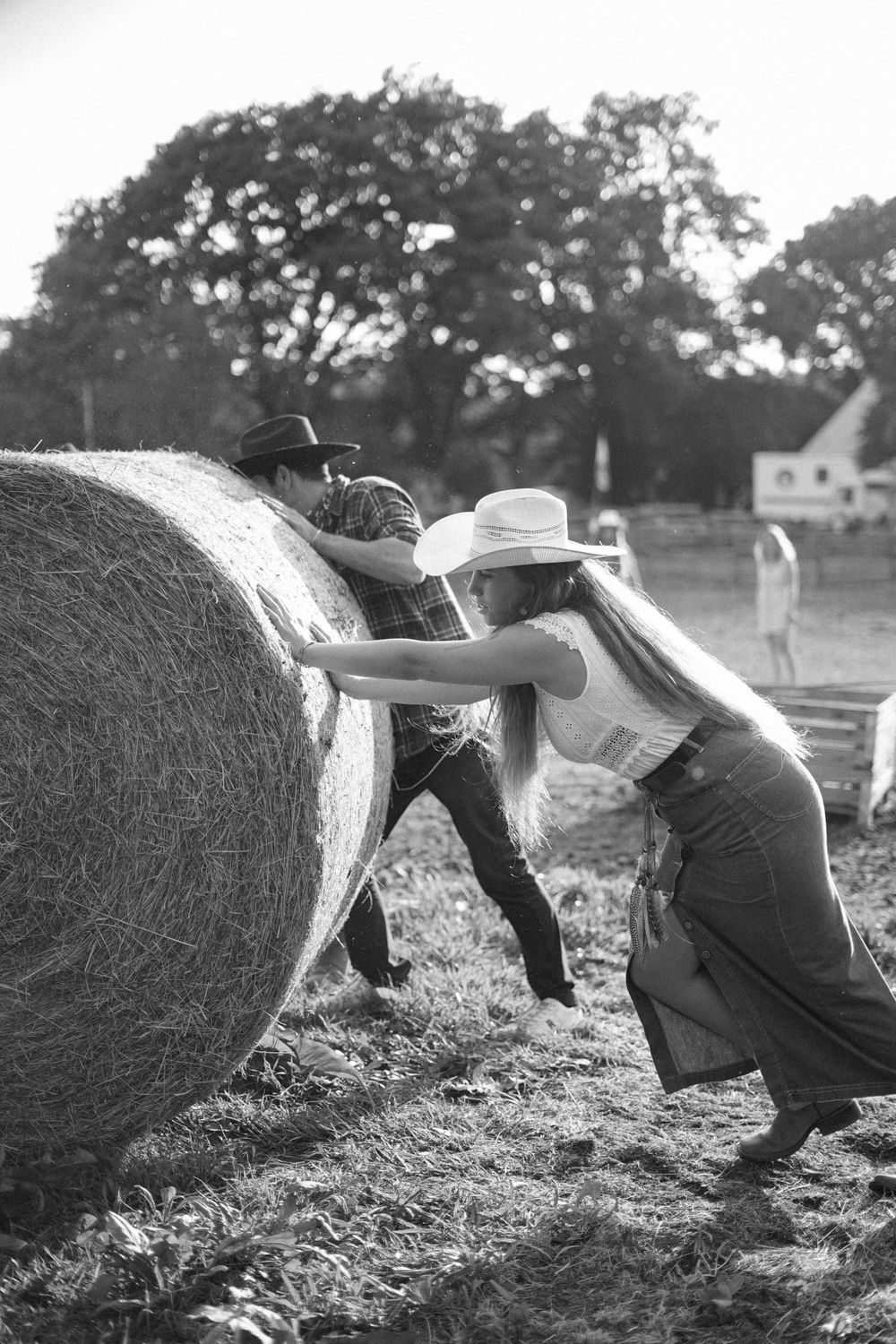 People in cowboy hats work together to push a large hay bale in a field.