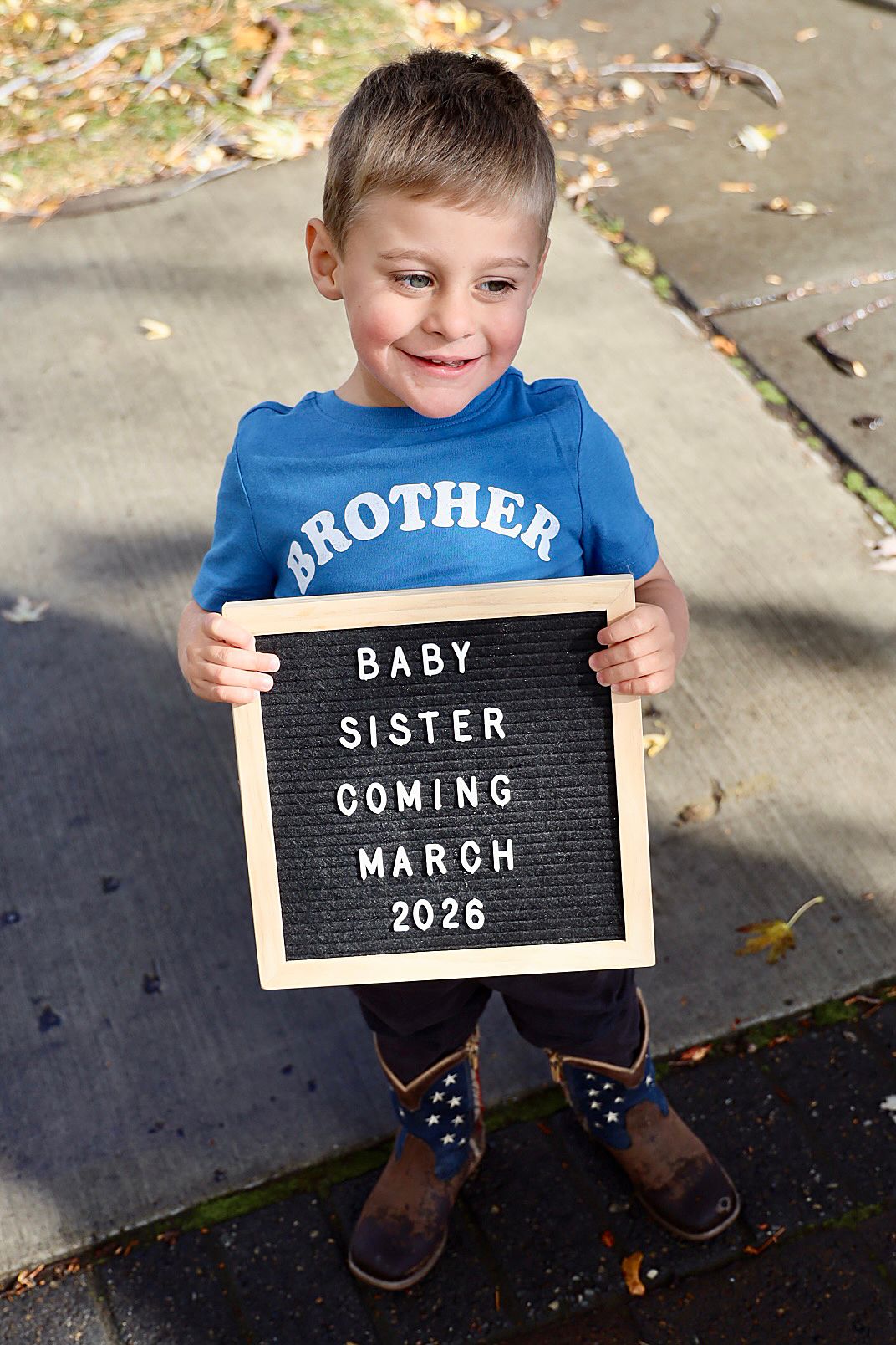 Young boy in Brother shirt holds letter board announcing Baby Sister Coming March 2026 in a sweet pregnancy reveal photo.