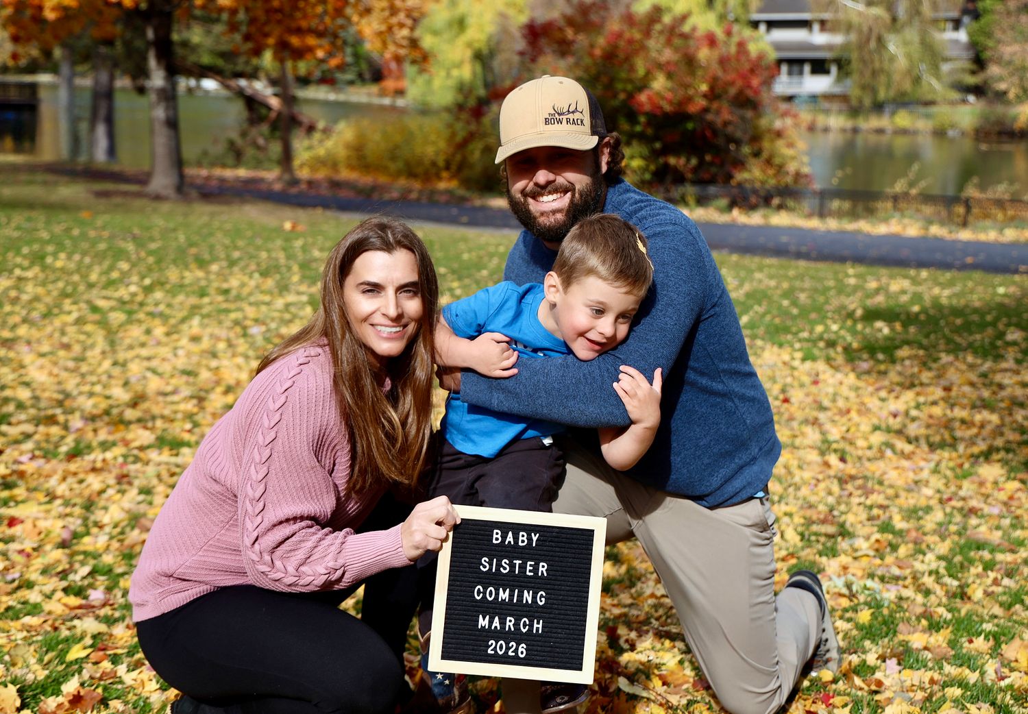 Family of three poses outdoors with a letter board announcing baby sister coming March 2024.