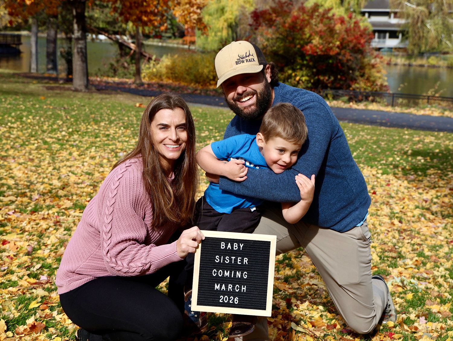 Family pregnancy announcement with letter board reading Baby Sister Coming March 2020, posed outdoors in autumn leaves.