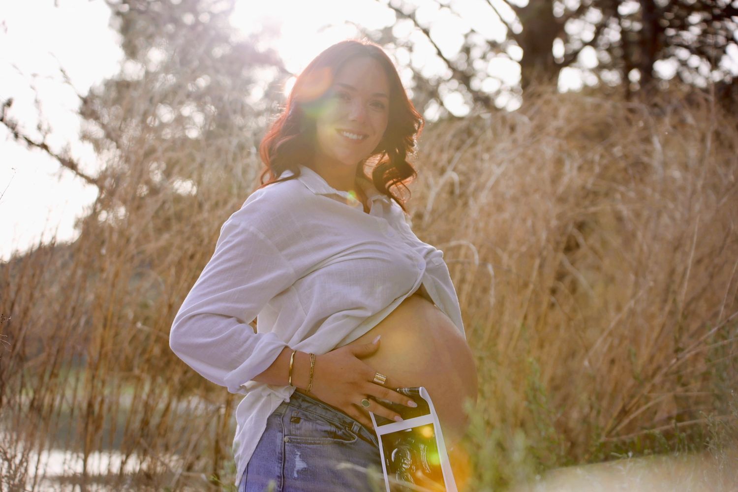 Pregnant woman holding ultrasound photo outdoors in golden sunlight, cradling baby bump in white shirt and denim shorts.