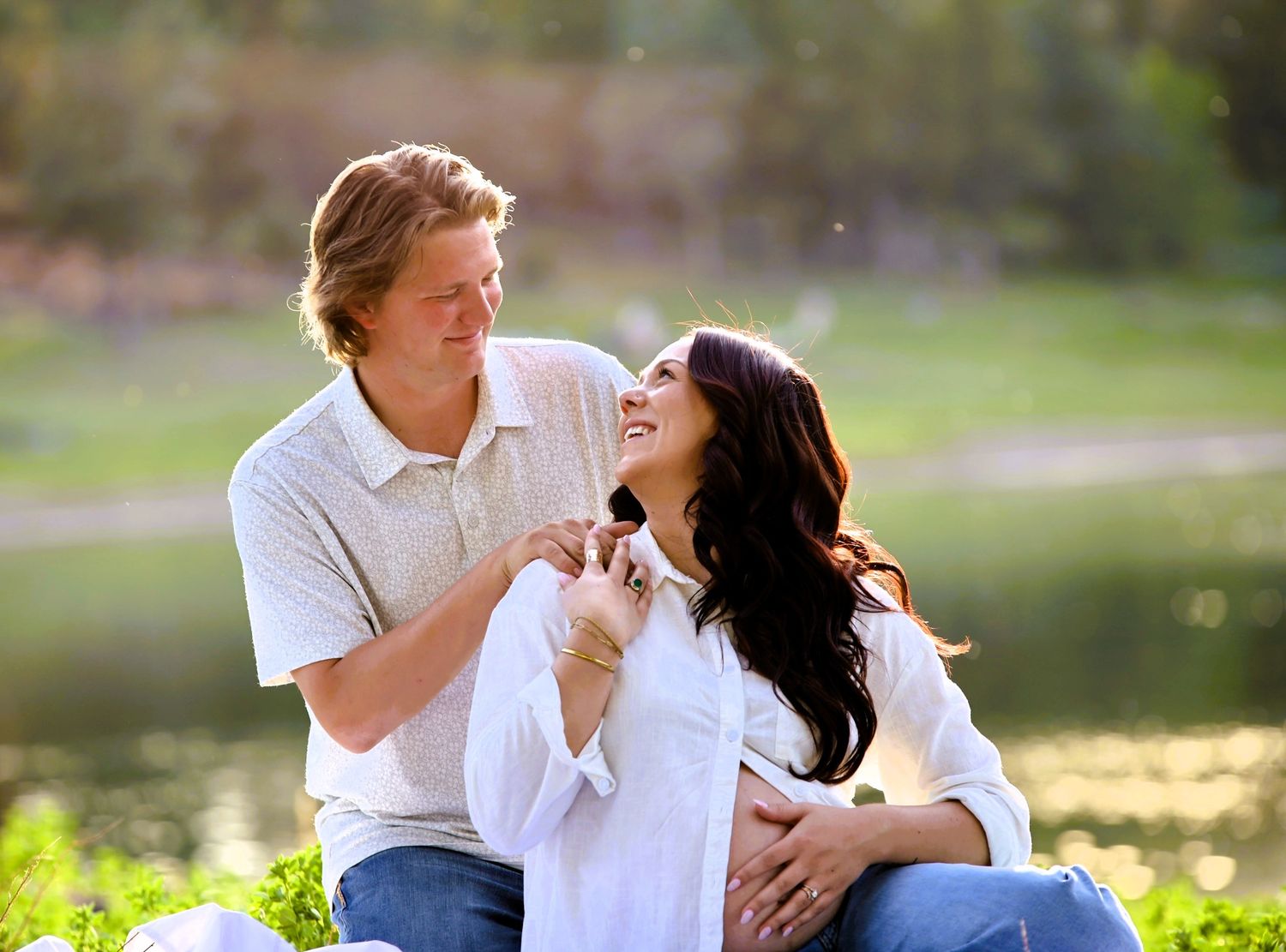 Happy couple sitting outdoors near a pond, woman in white shirt smiling up at blond man in sunny park setting.