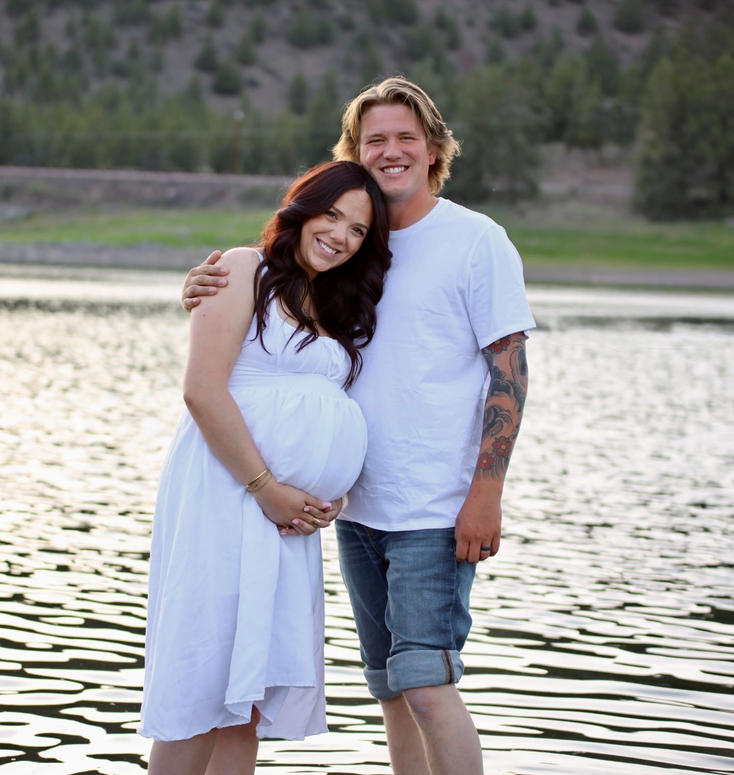 Pregnant woman in white dress and man in white t-shirt smiling by a lake during maternity photo shoot.
