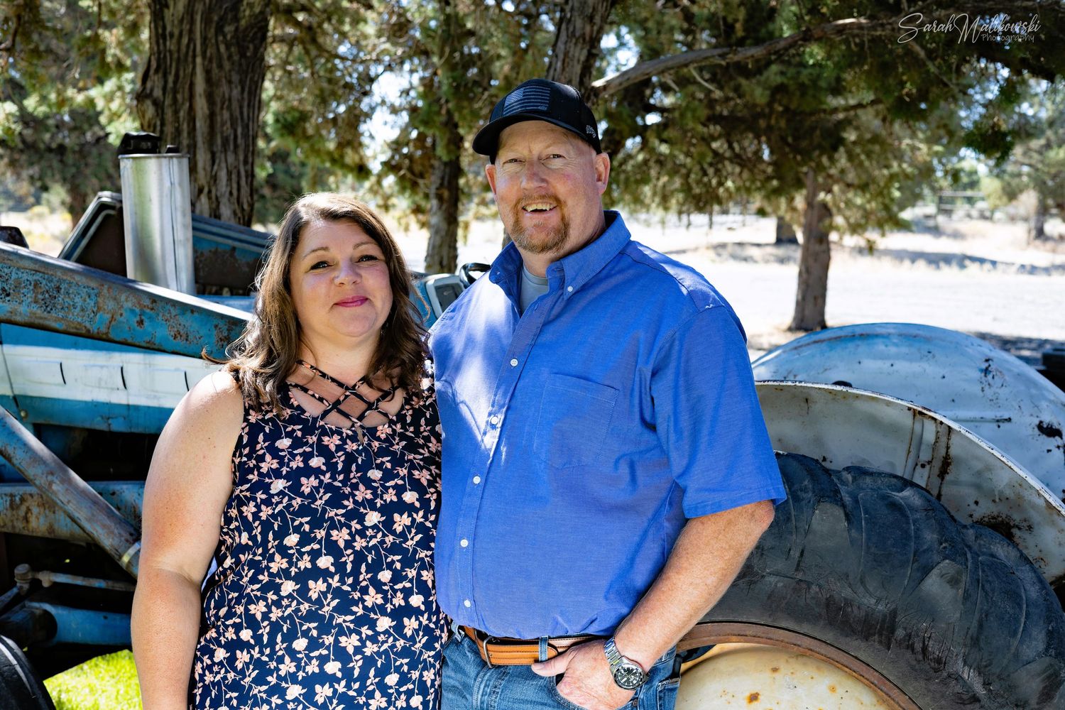 A couple in casual attire poses together in front of farming equipment outdoors.