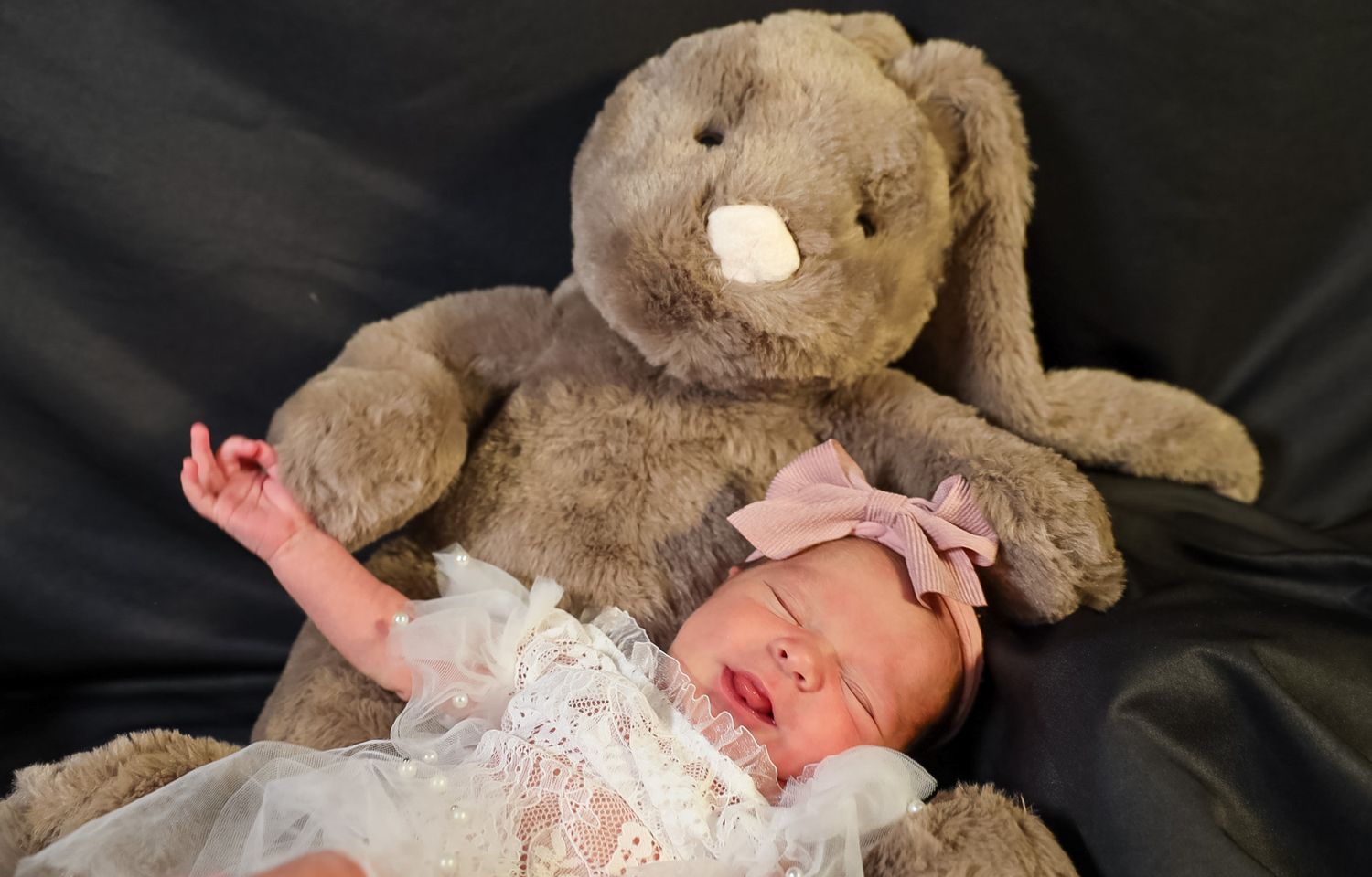 Newborn baby in white lace outfit with pink bow headband sleeping beside a large gray stuffed bunny on black background.