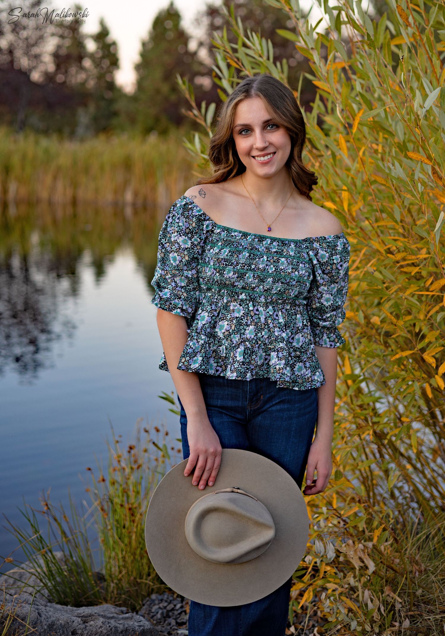 Outdoor portrait series by a lake featuring floral top and wide-brimmed hat.