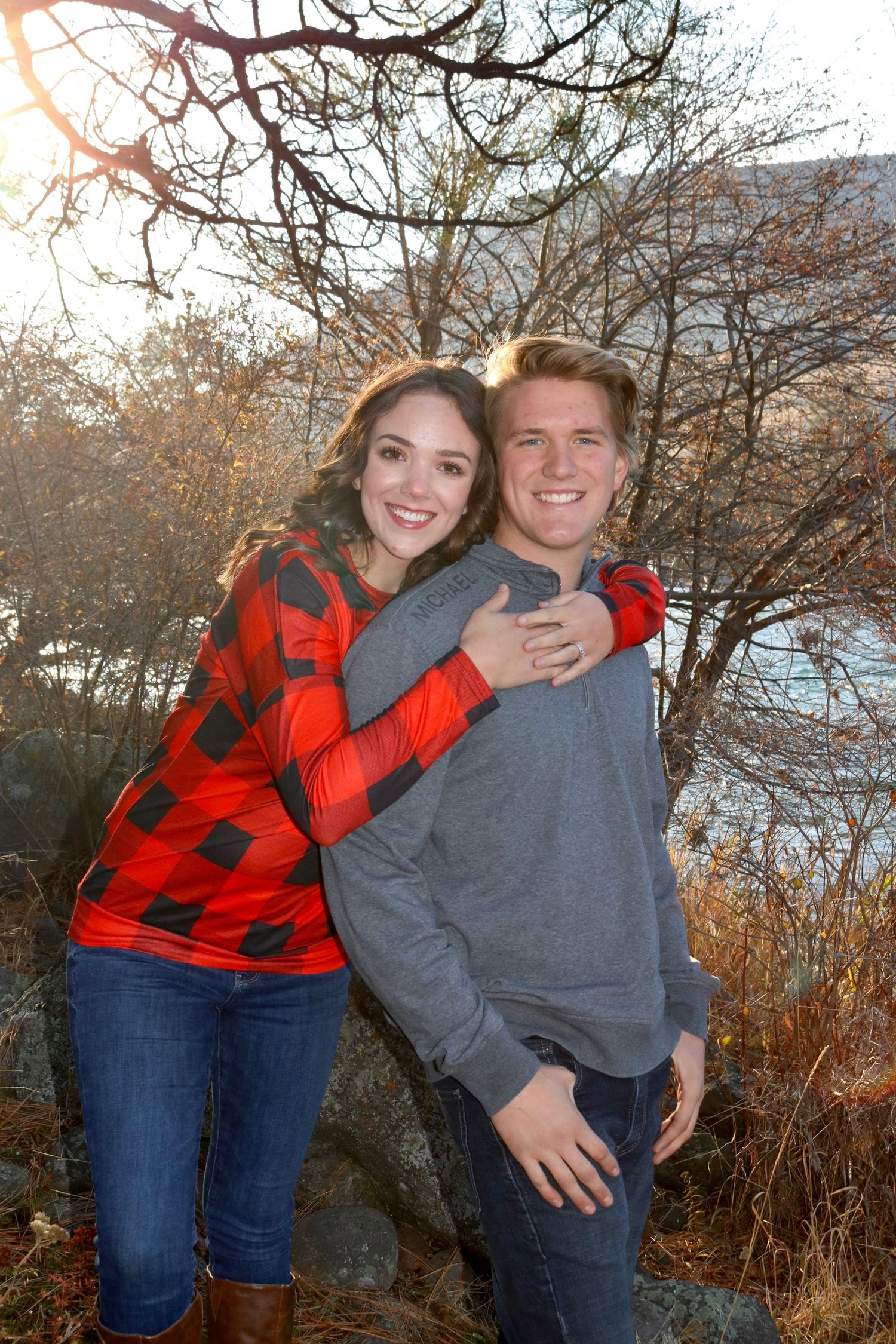 A couple in casual winter clothing embraces outdoors among bare trees.