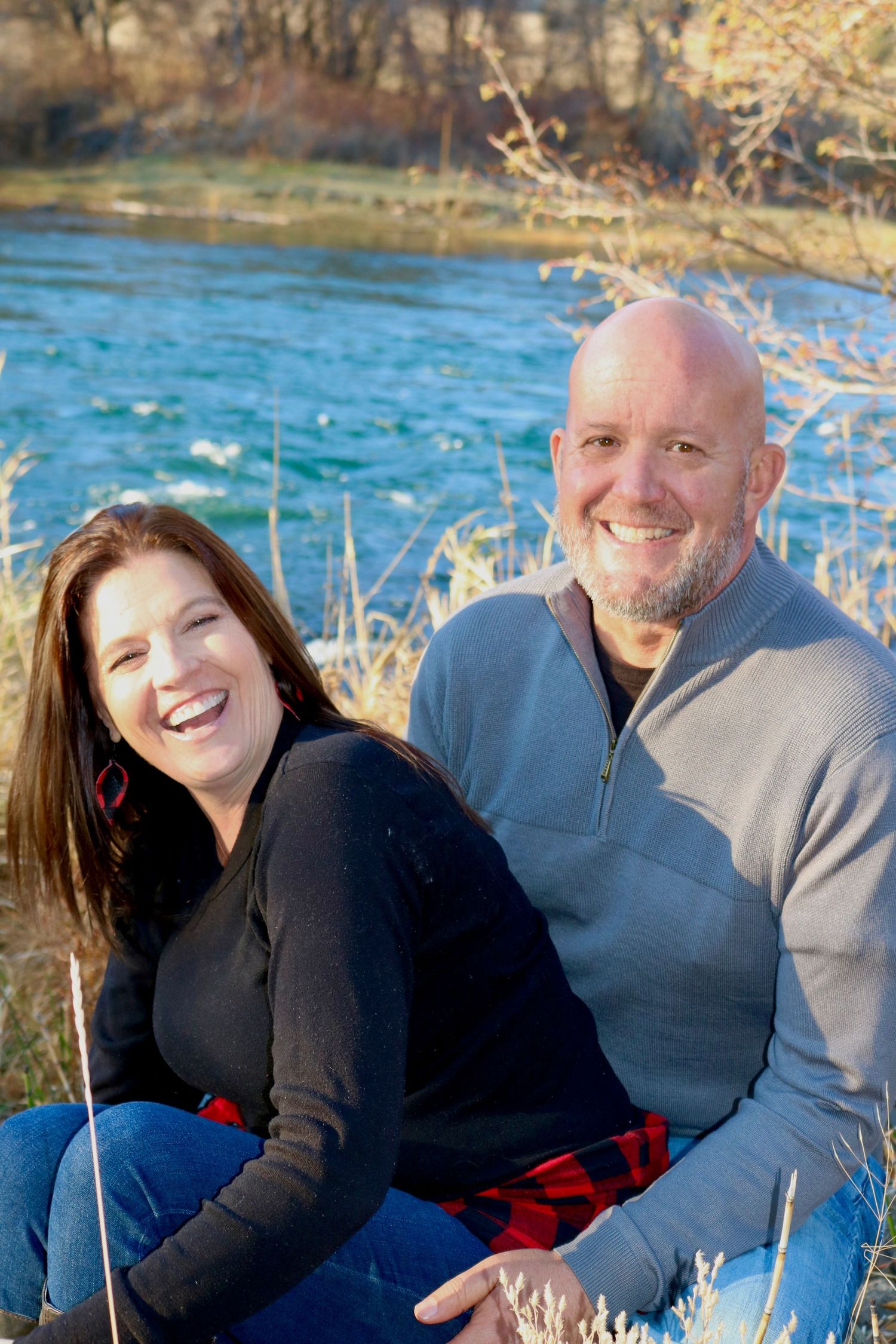 A couple sits together by a blue river on a sunny winter day.