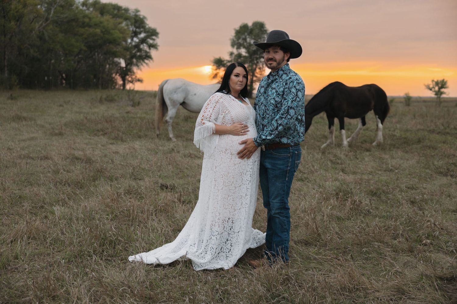 Pregnant couple poses at sunset in a field with horses in Garrison, ND. woman in white lace gown and man in cowboy hat very western attire