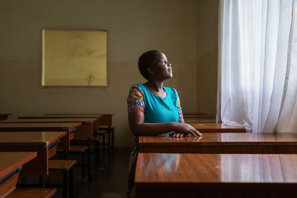 A hopeful, happy orphan student sits a desk facing a lit window at a children's home in Malawi. orphan-school-Malawi