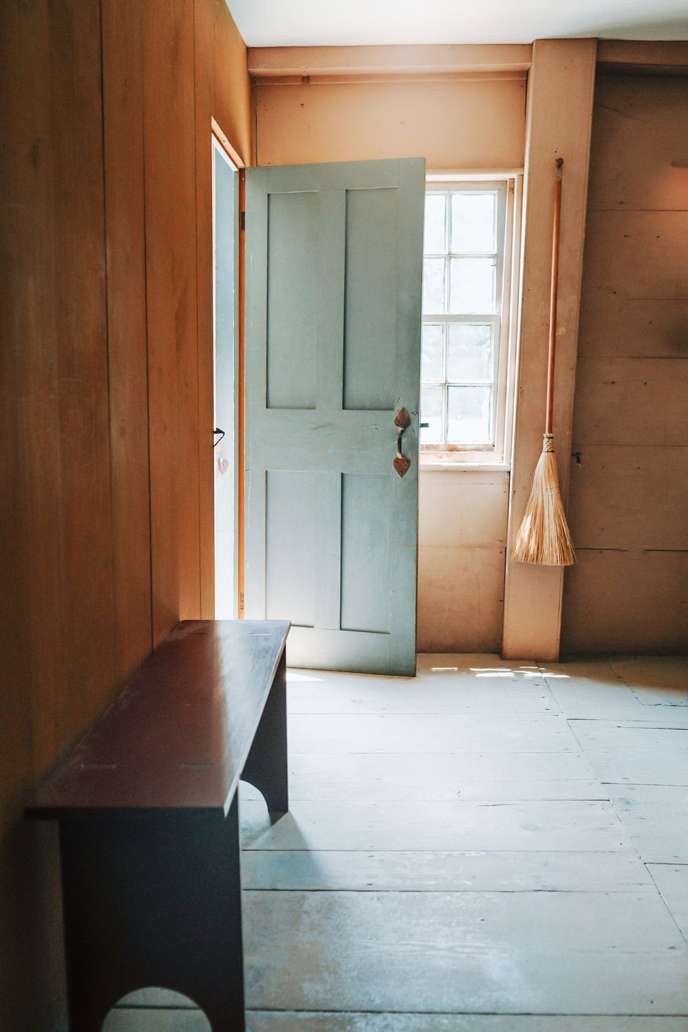 A mint colored door opens into a plain historical wood paneled room with a broom leaning against a post at Sturbridge Village in MA.