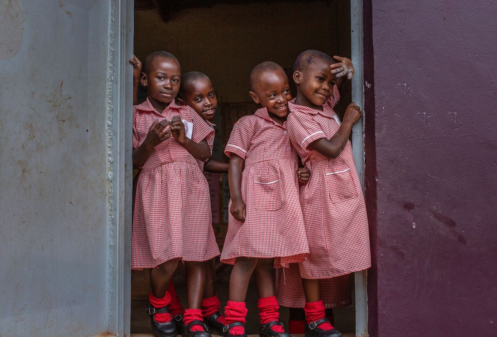 Orphan children wearing red gingham dresses stand in a doorway with gray and purple walls in a school house in Uganda. orphans-Uganda