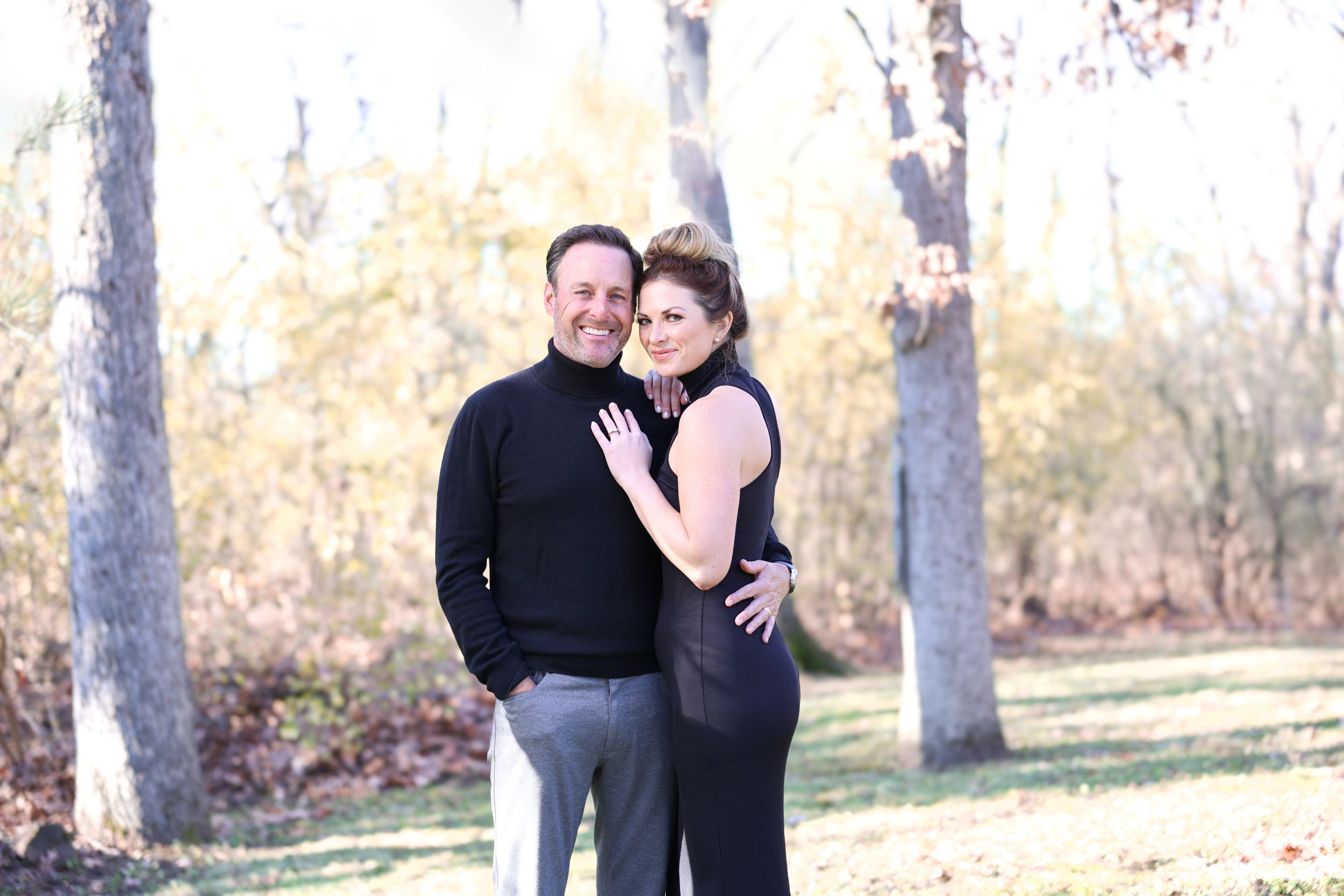 A couple poses together in an outdoor autumn setting with trees and fallen leaves in the background.