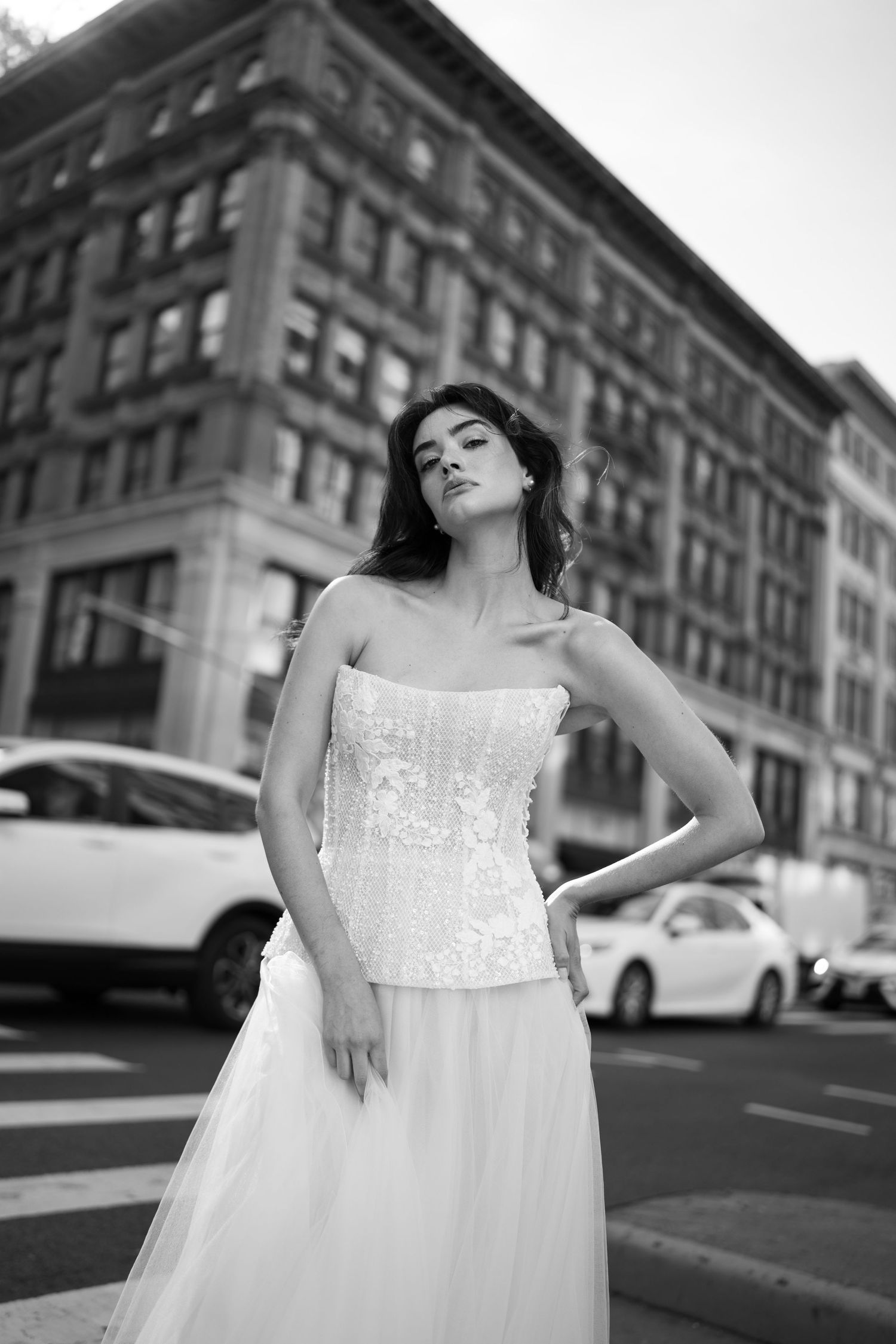 A black and white fashion photograph featuring a strapless wedding dress with corset bodice against an urban backdrop.
