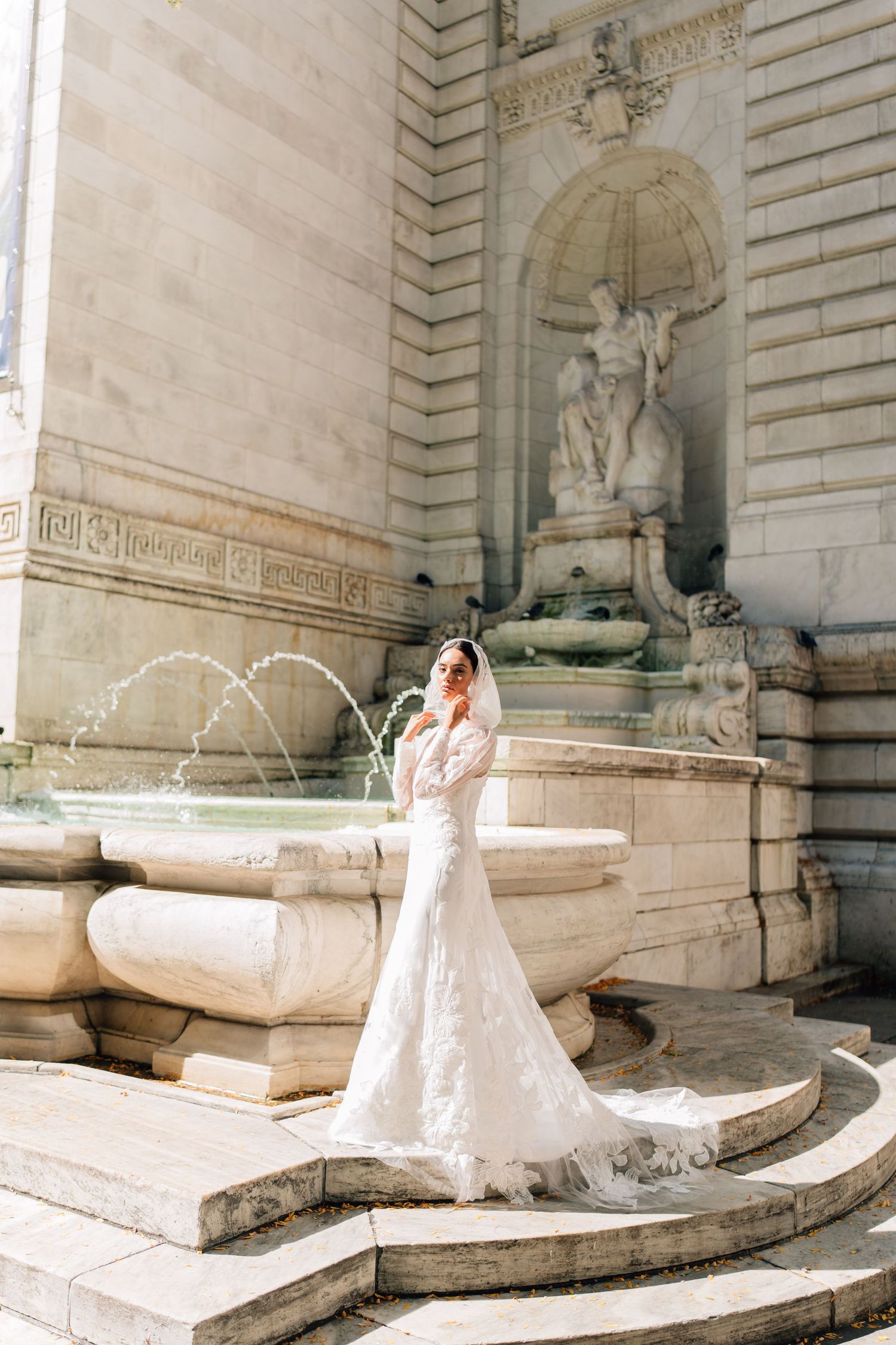 A person in a white wedding dress sits near stone benches and columns in an ornate architectural setting.