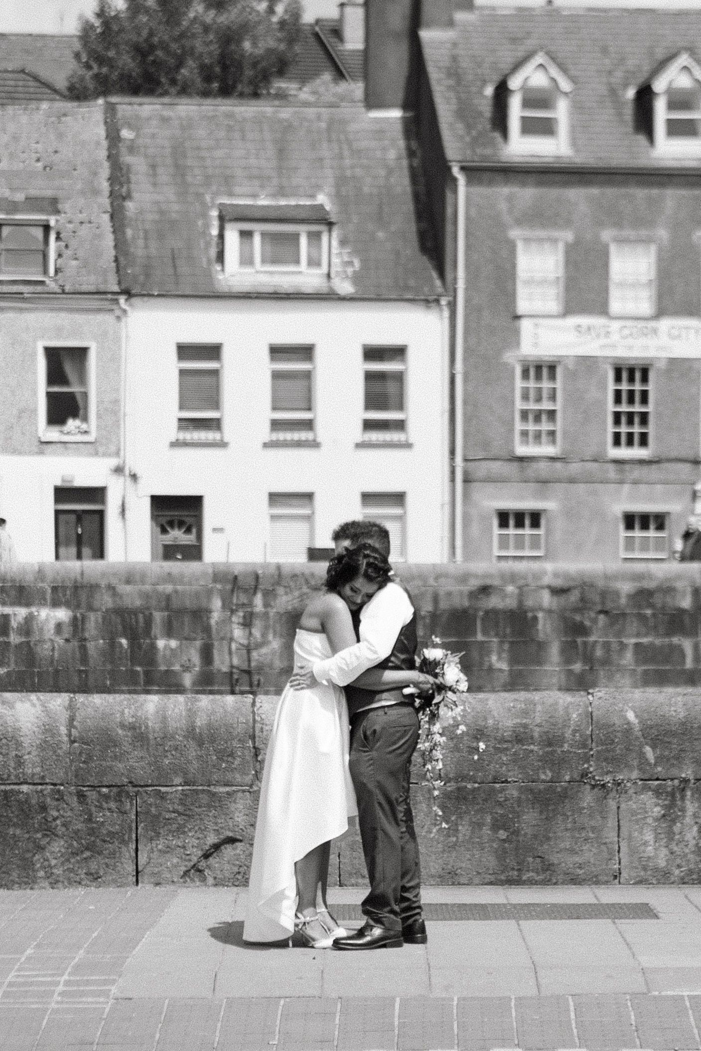 Black and white photo of a couple embracing on a street with historic buildings in the background.