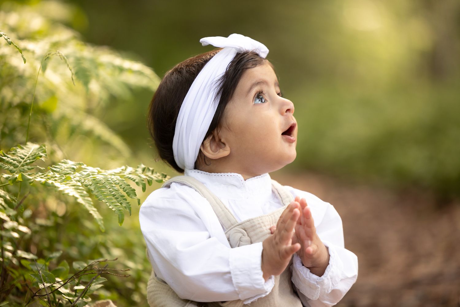 Young child in white dress and headband looking up in wonder amid green foliage.
