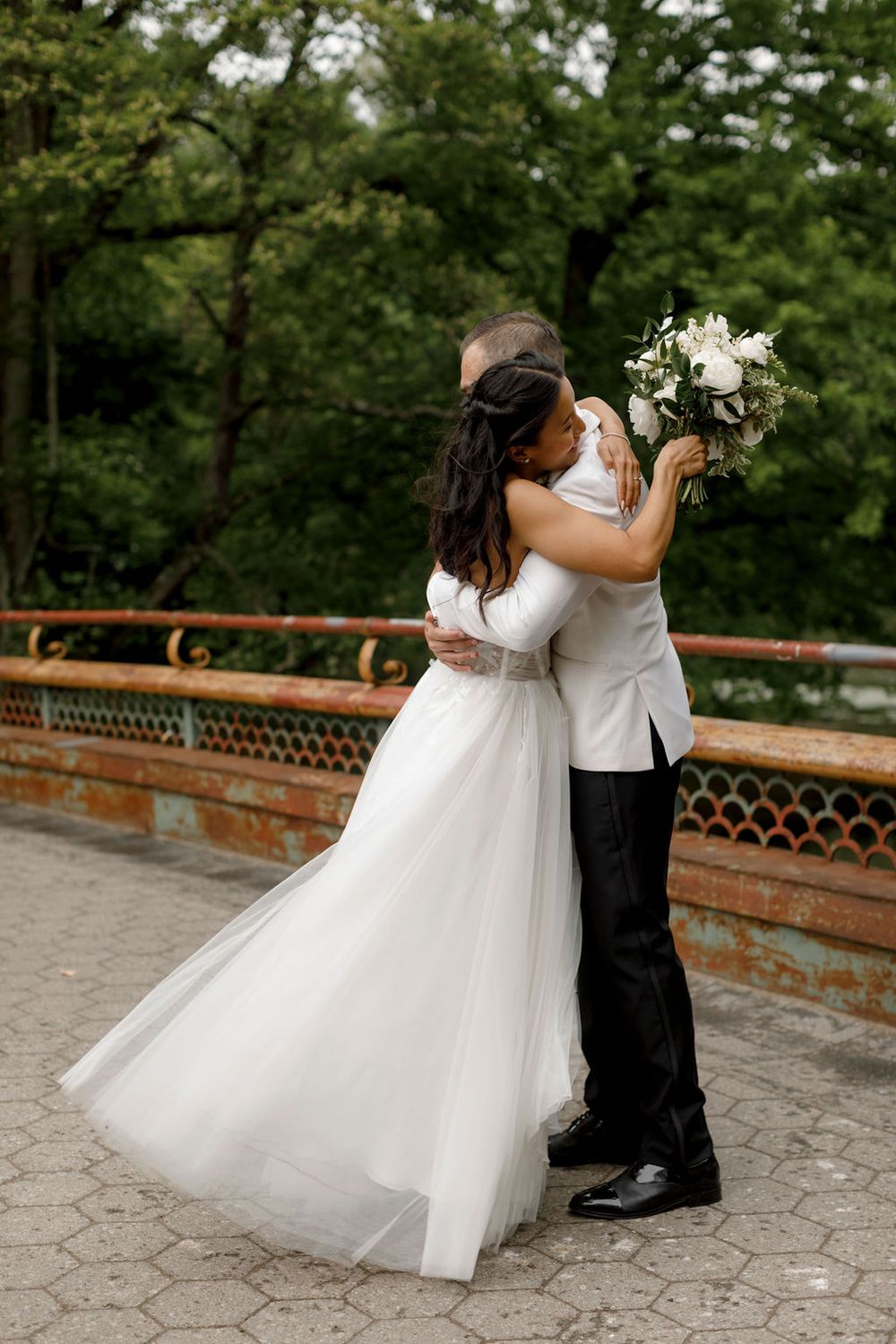 Newlyweds embracing on stone bridge with bride in flowing white gown holding bouquet.