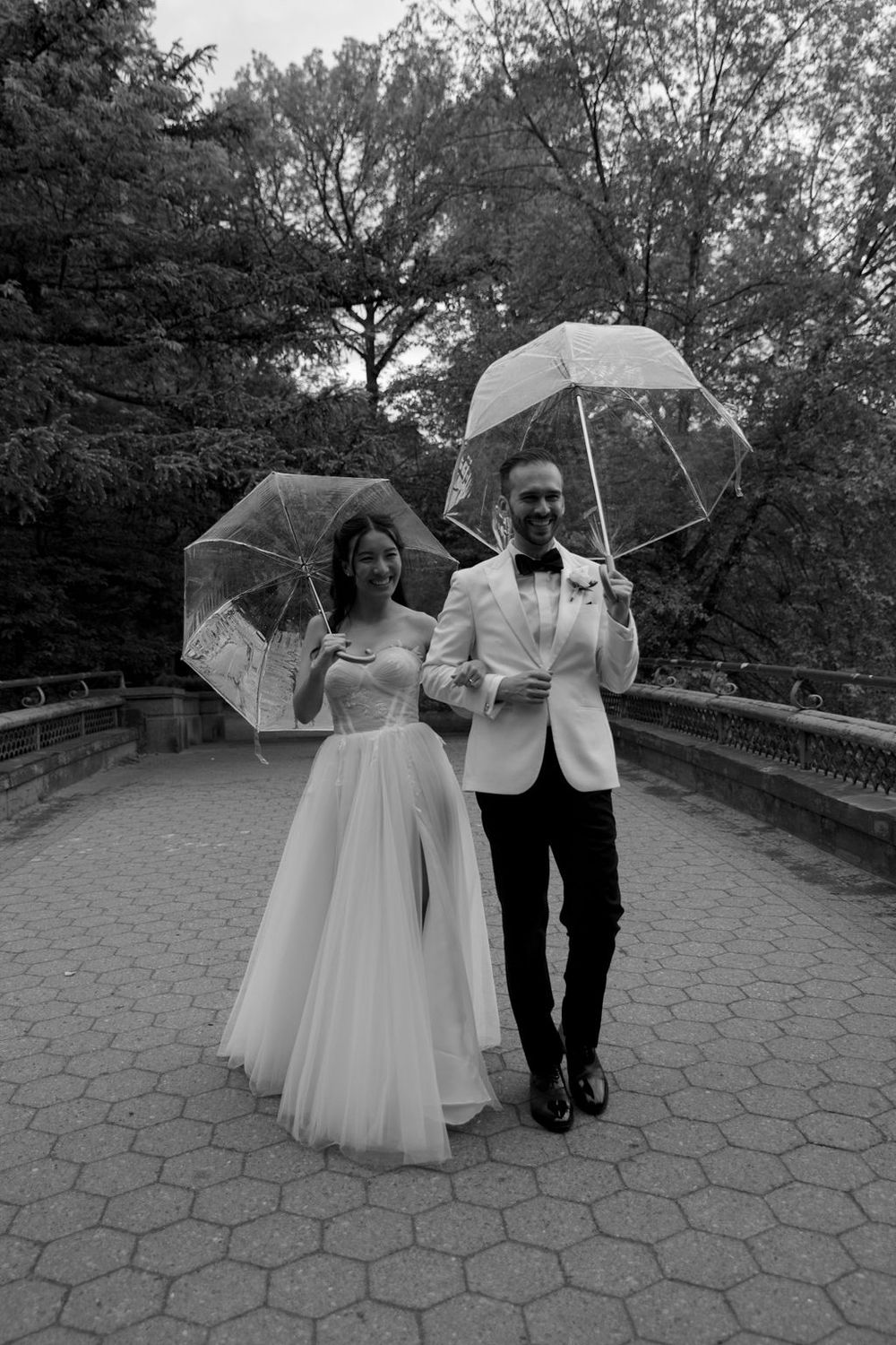 Black and white photo of newlyweds with clear umbrellas on cobblestone bridge.